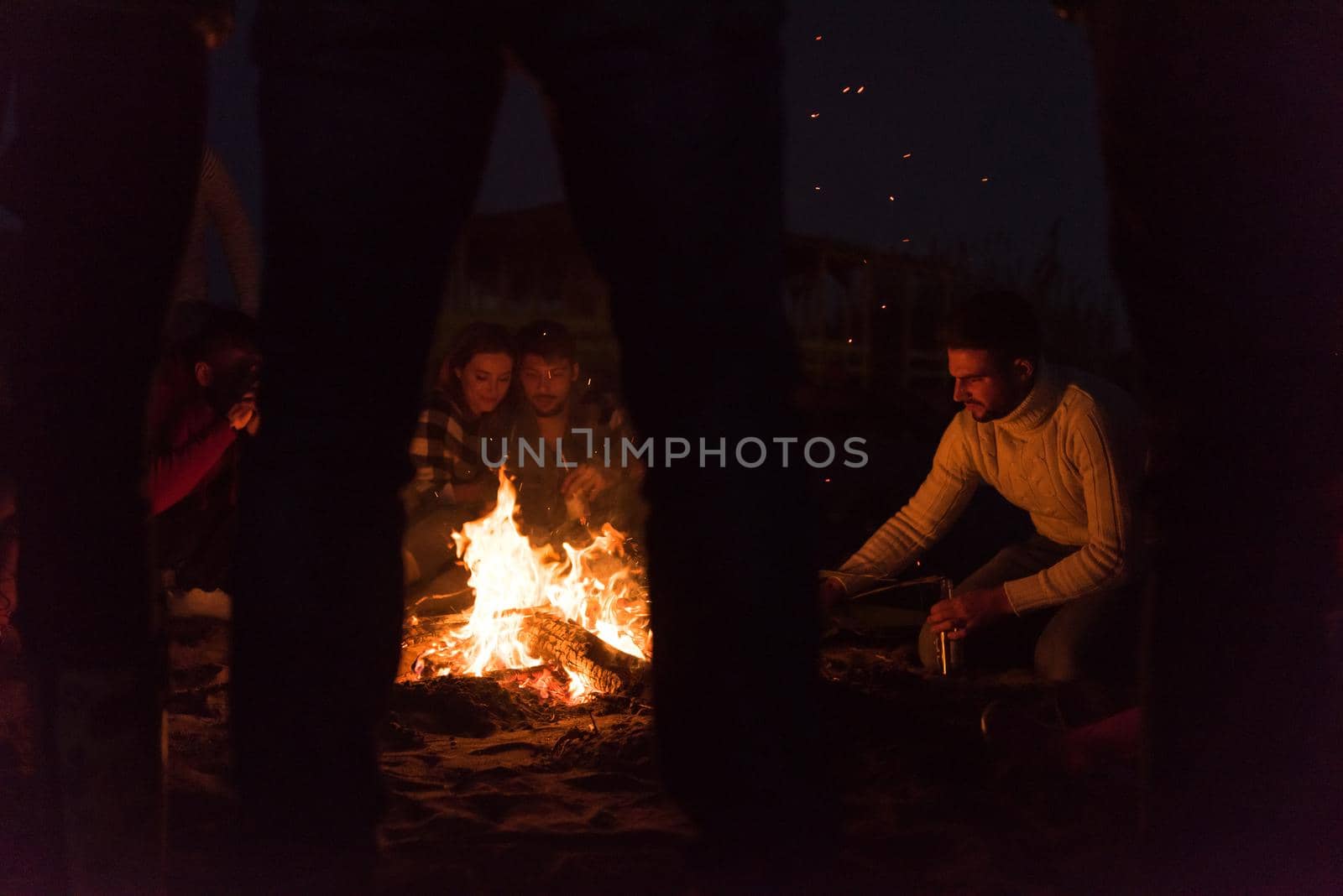 Happy Carefree Young Friends Having Fun And Drinking Beer By Bonefire On The Beach As The Sun Begins To Set