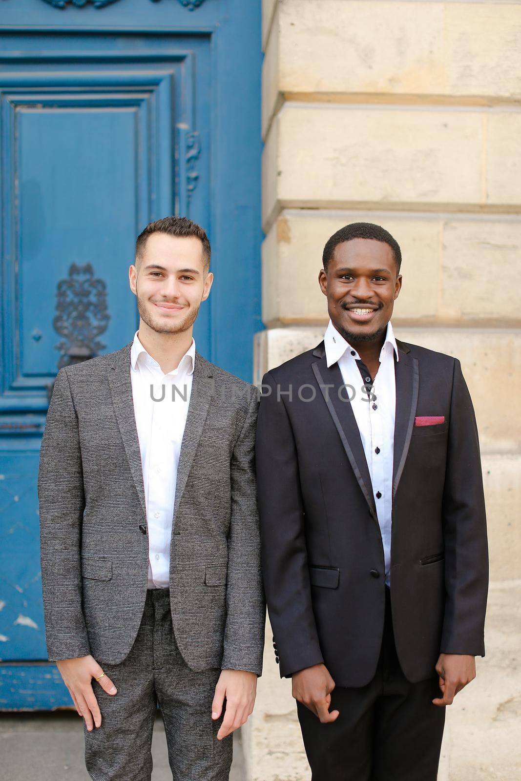 Two men, caucasian and afro american, wearing suits standing near building. by sisterspro