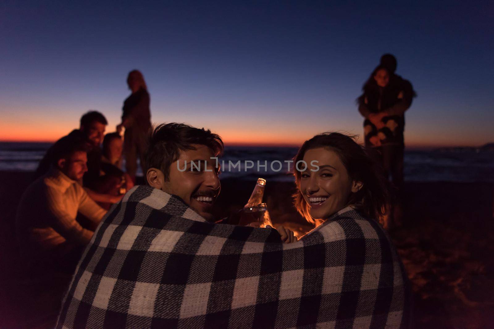 Young Couple enjoying with friends Around Campfire on The Beach At sunset drinking beer