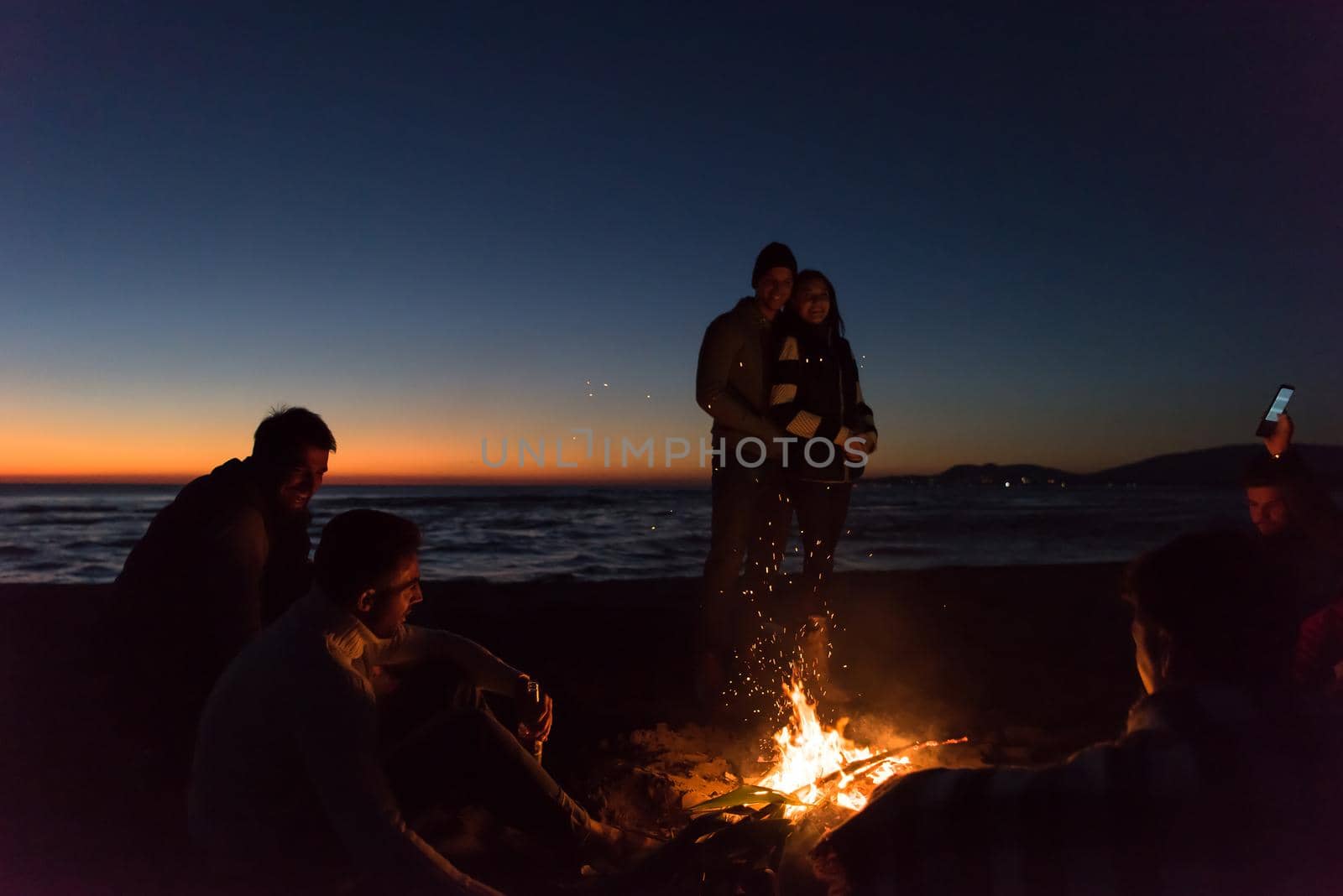 Happy Carefree Young Friends Having Fun And Drinking Beer By Bonefire On The Beach As The Sun Begins To Set