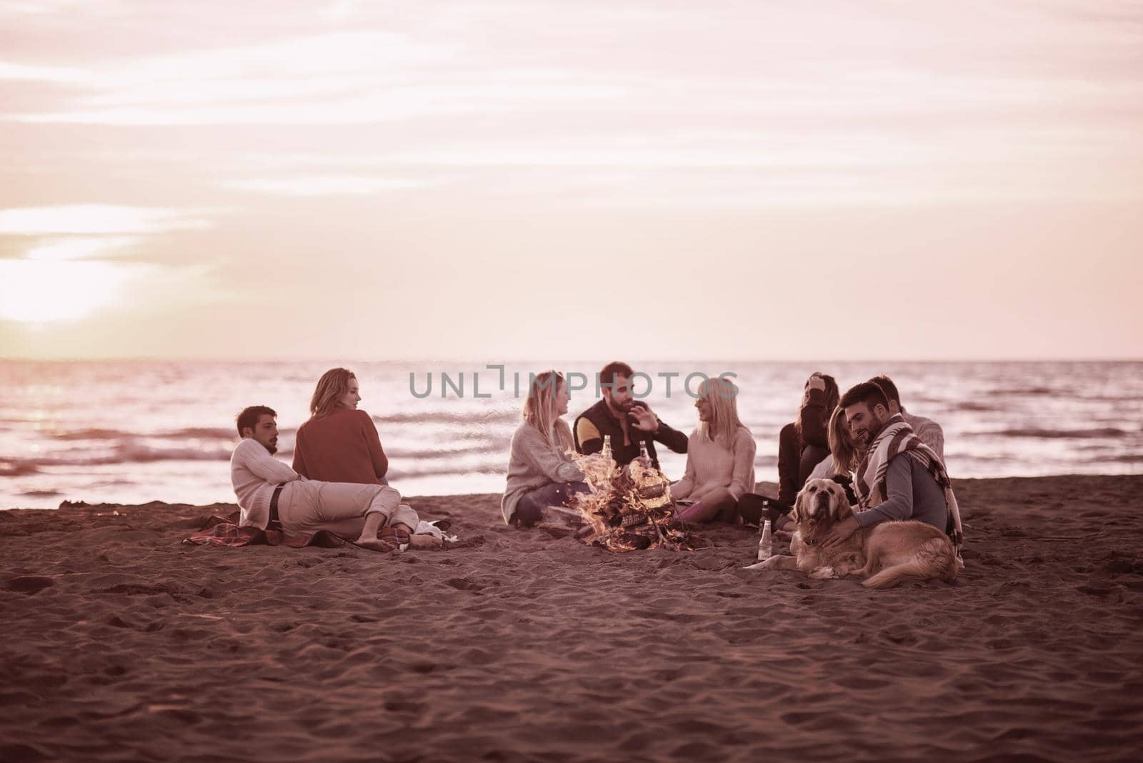 Happy Carefree Young Friends Having Fun And Drinking Beer By Bonefire On The Beach As The Sun Begins To Set filter