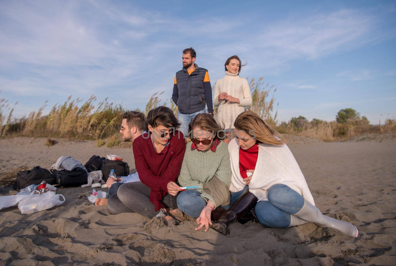 Happy Carefree Young Friends Having Fun And Drinking Beer By Bonefire On The Beach As The Sun Begins To Set