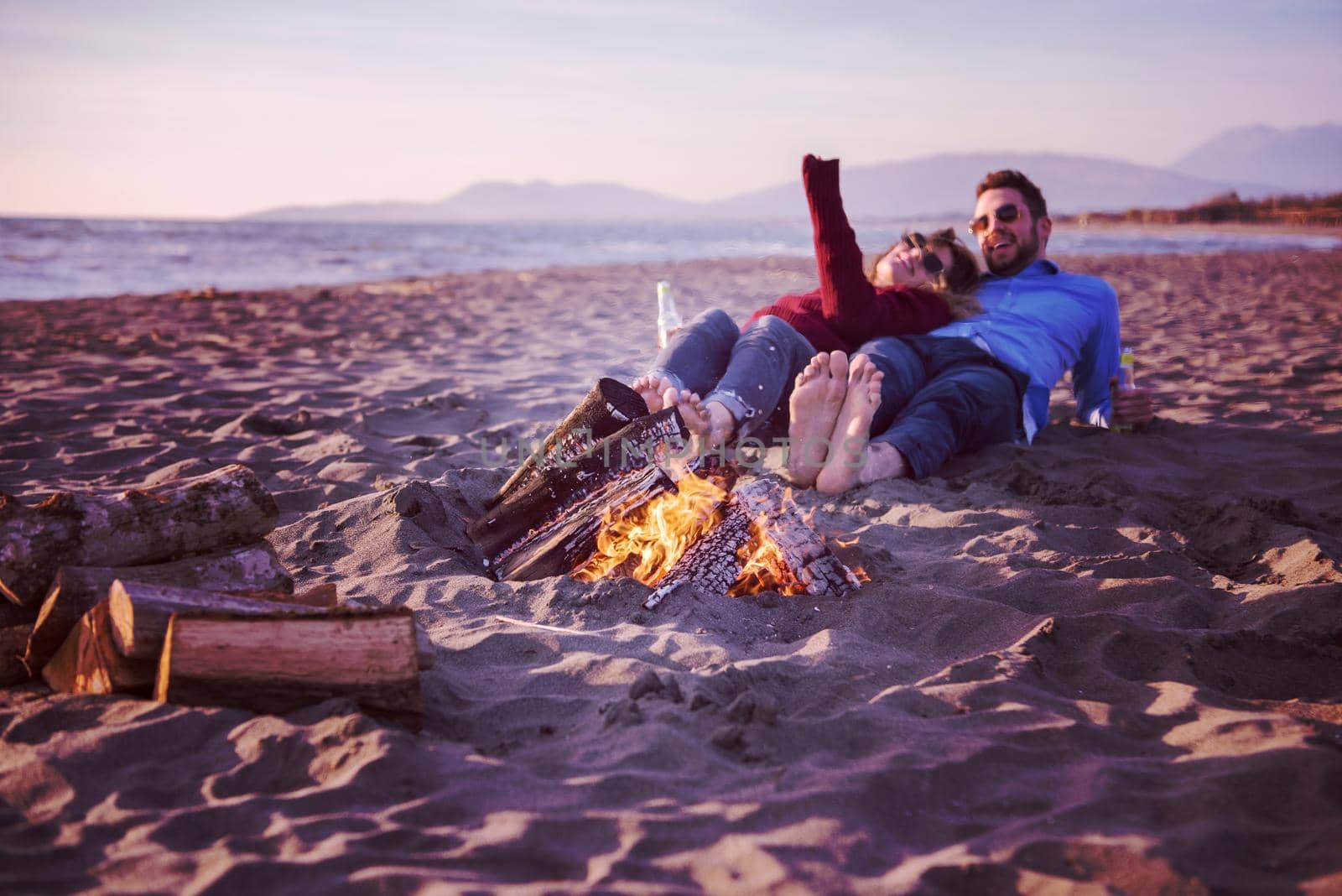 Young Couple Relaxing By The Fire, Drinking A Beer Or A Drink From The Bottle on the beach at autumn day filter