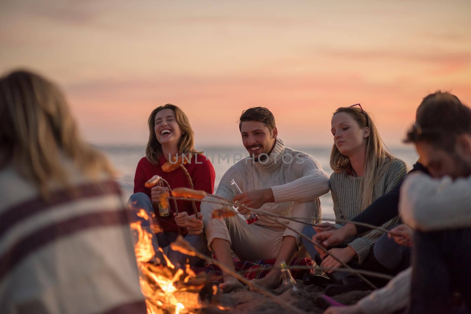 Group of young friends sitting by the fire at autumn beach, grilling sausages and drinking beer, talking and having fun