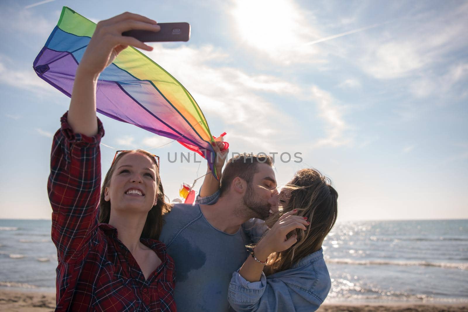 group of young friends making selfie while playing with kite on a beach during sunny autumn day