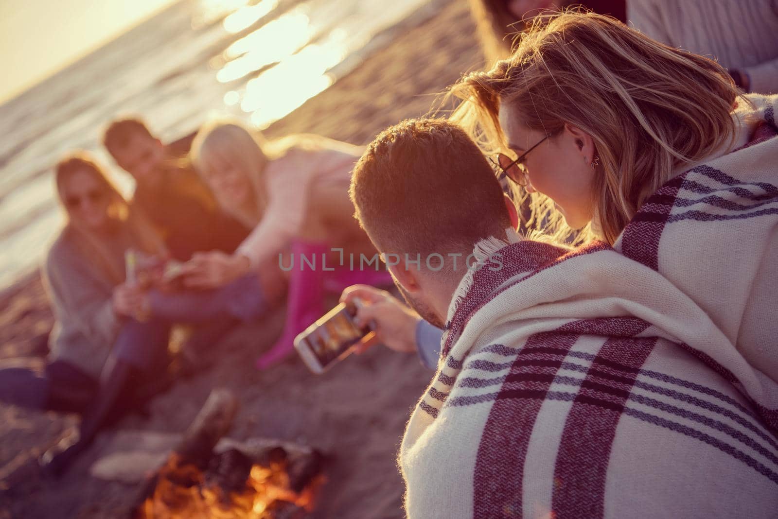 Happy Carefree Young Friends Having Fun And Drinking Beer By Bonefire On The Beach As The Sun Begins To Set filter