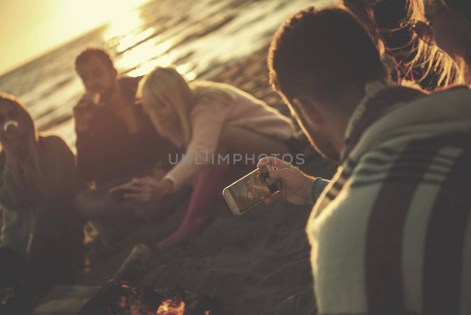 Happy Carefree Young Friends Having Fun And Drinking Beer By Bonefire On The Beach As The Sun Begins To Set filter