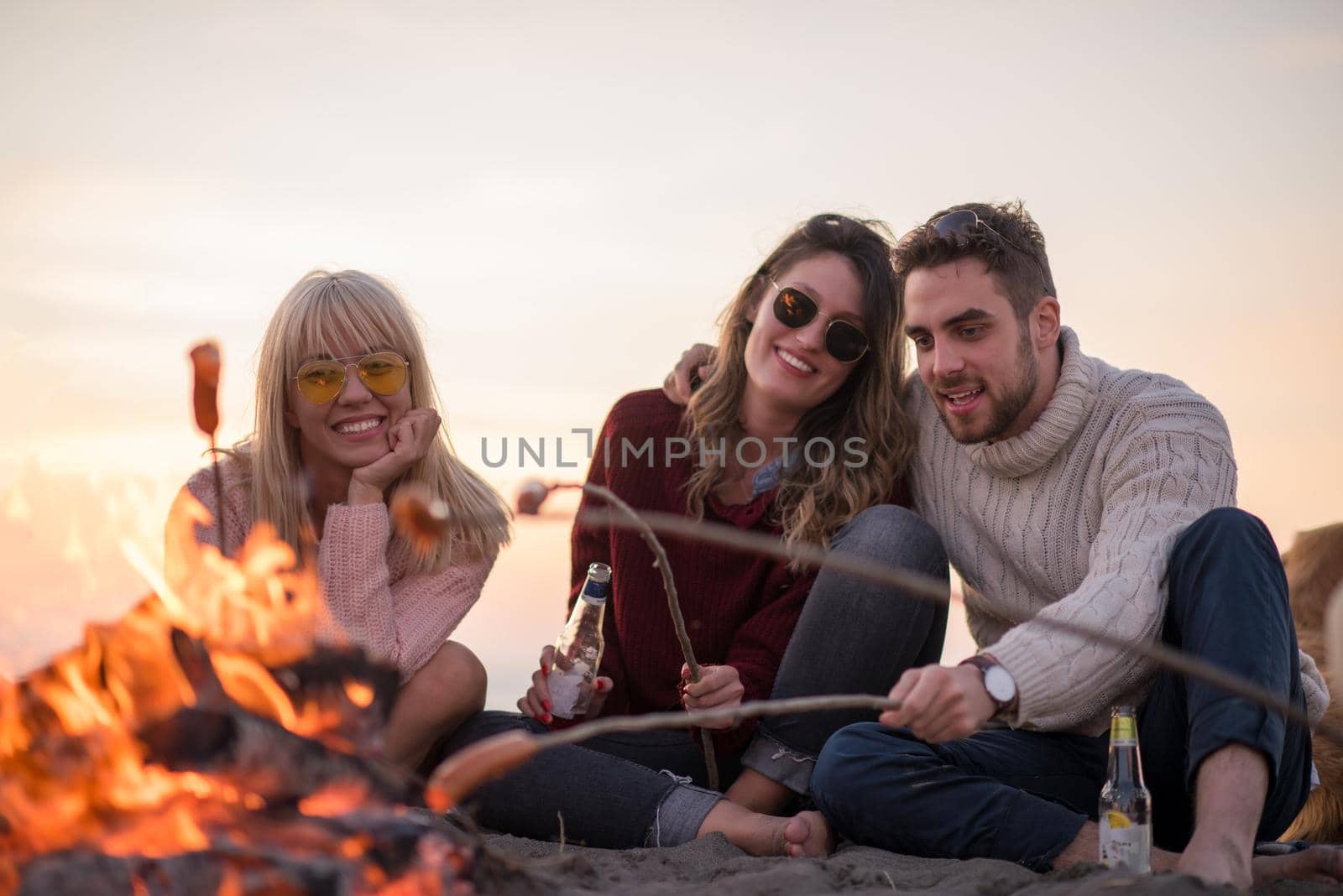 Group of young friends sitting by the fire at autumn beach, grilling sausages and drinking beer, talking and having fun