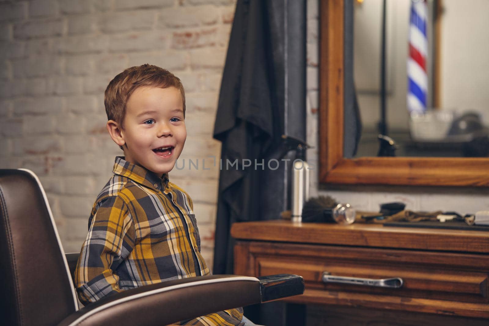 Portrait of a stylish little boy dressed in shirt and jeans sitting in a chair against the barber's workplace, he smiles and posing before camera