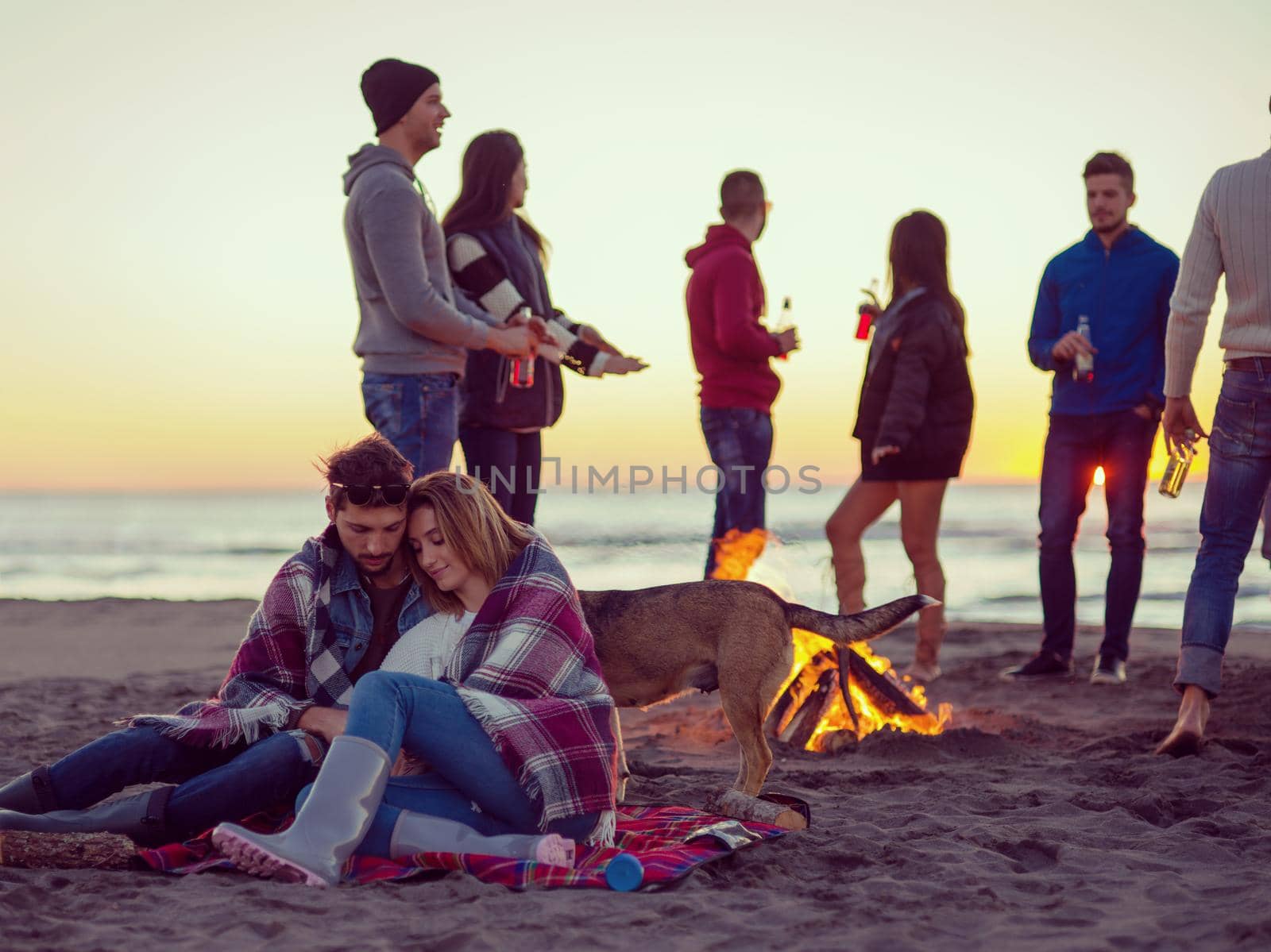 Young Couple Sitting with friends Around Campfire on The Beach At sunset drinking beer colored filter