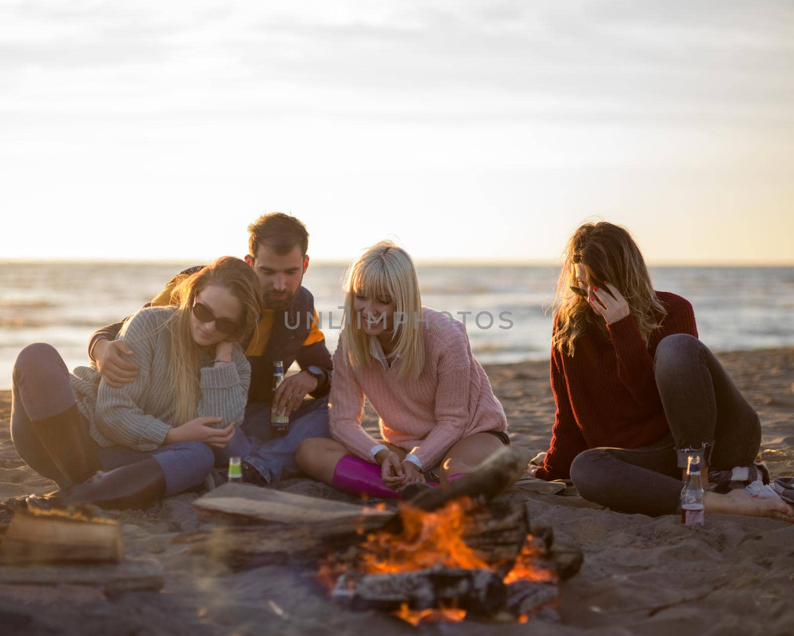 Happy Carefree Young Friends Having Fun And Drinking Beer By Bonefire On The Beach As The Sun Begins To Set