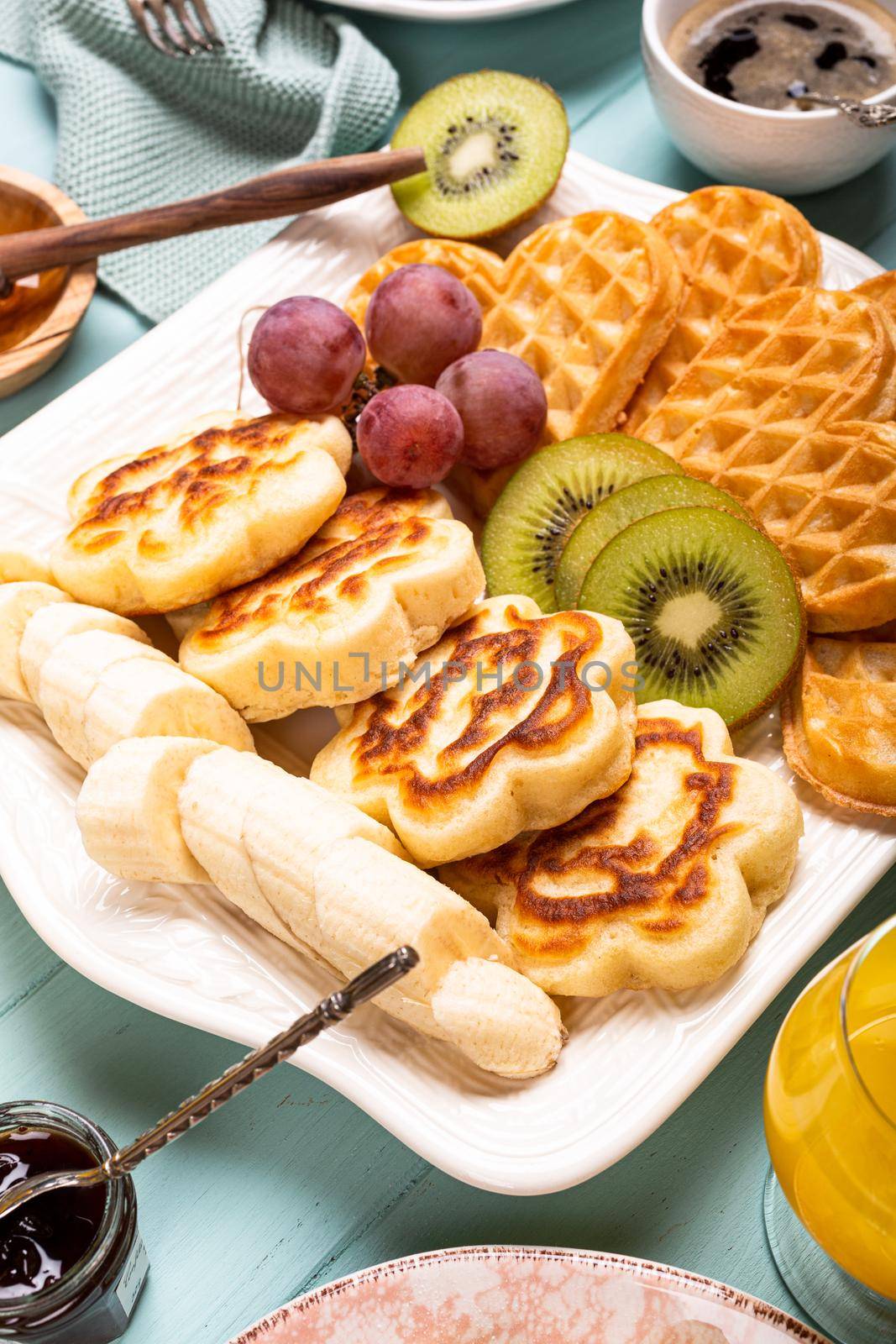 Fresh hot flowers pancakes with waffles hearts, berry jam and fruits on turquoise background. Healthy food concept.