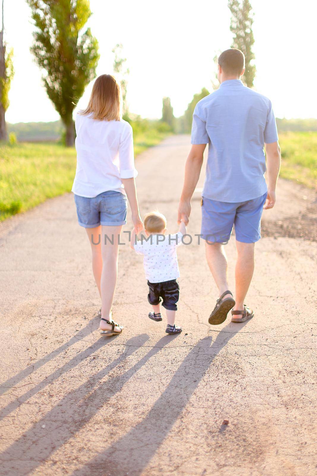 Back view of young mother and father walking on road with little baby, sunshine weather. Concept of parents and children, resting on nature.
