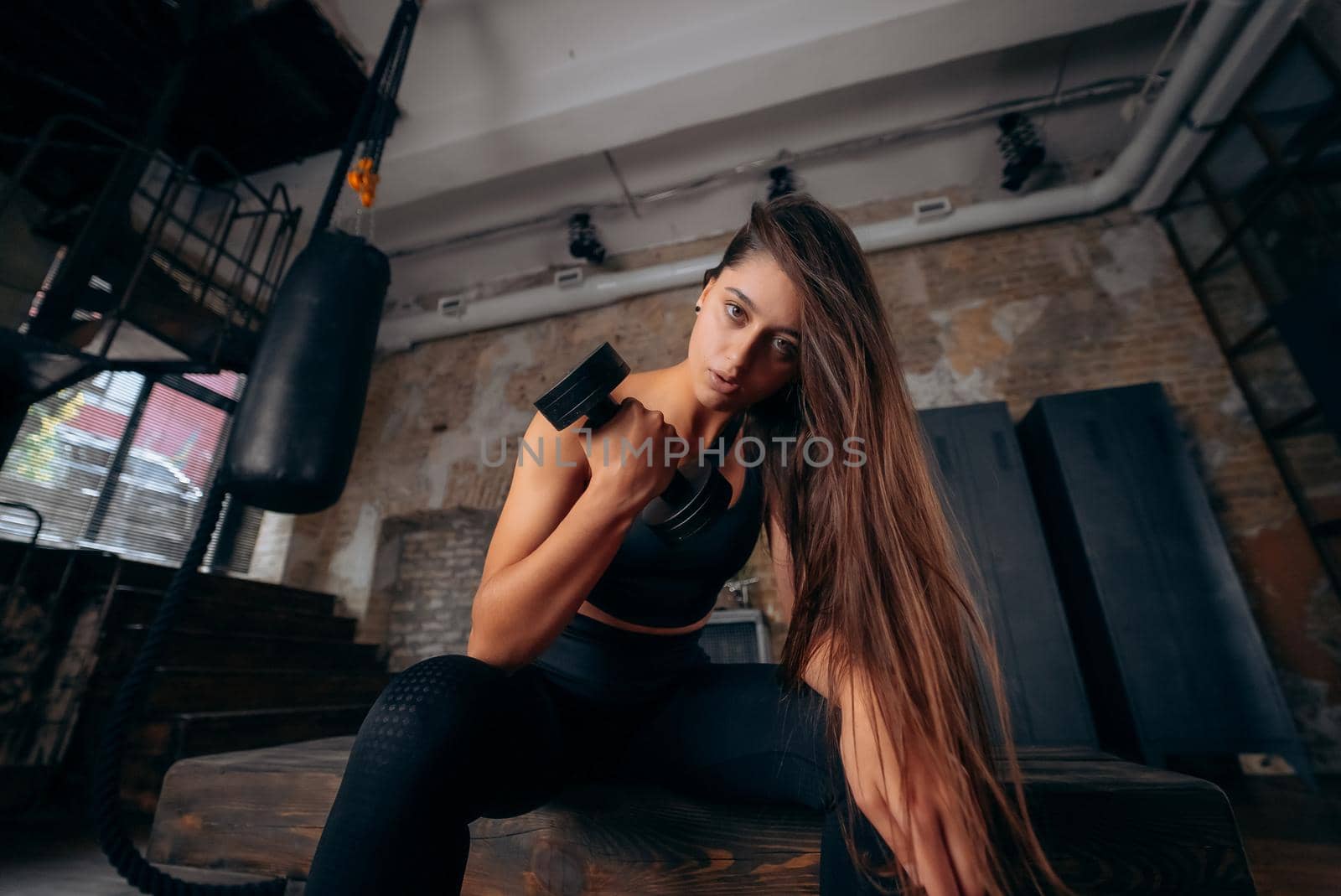 Young woman pumps up the muscles by one arm lifting dumbbell. Evening exercise on bench in sport gym.