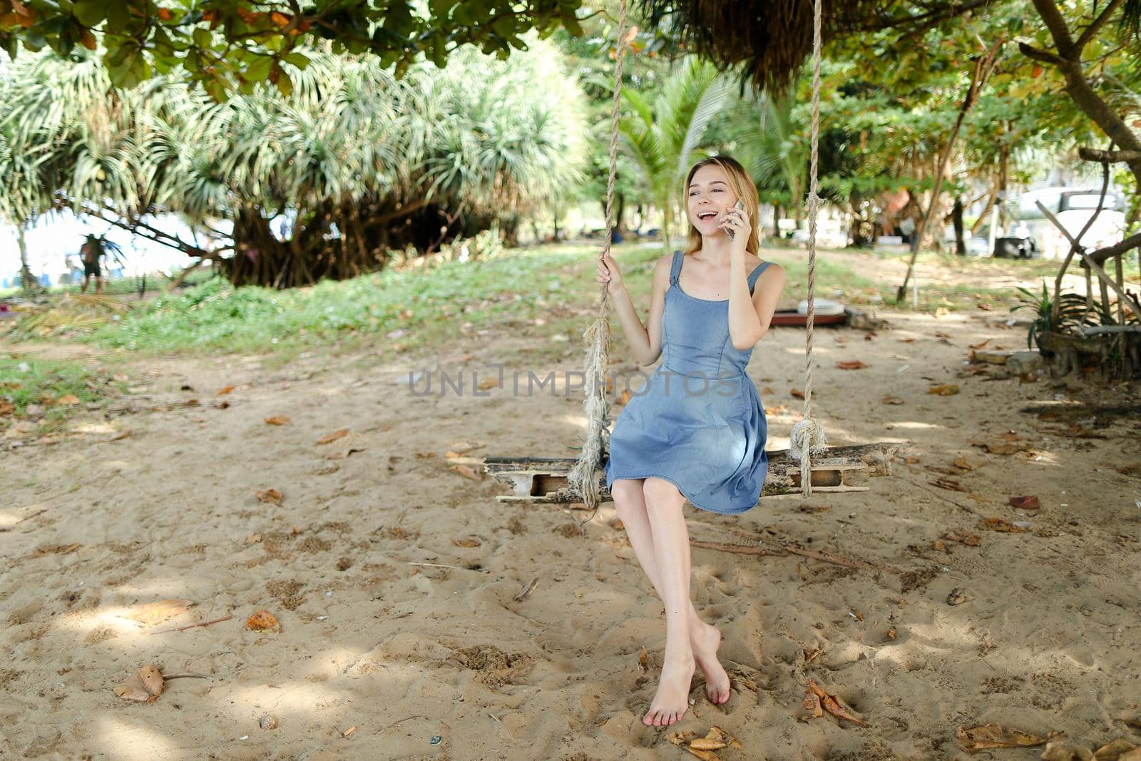 Young european woman riding swing and speaking by smatrphone, sand and tree in background. by sisterspro