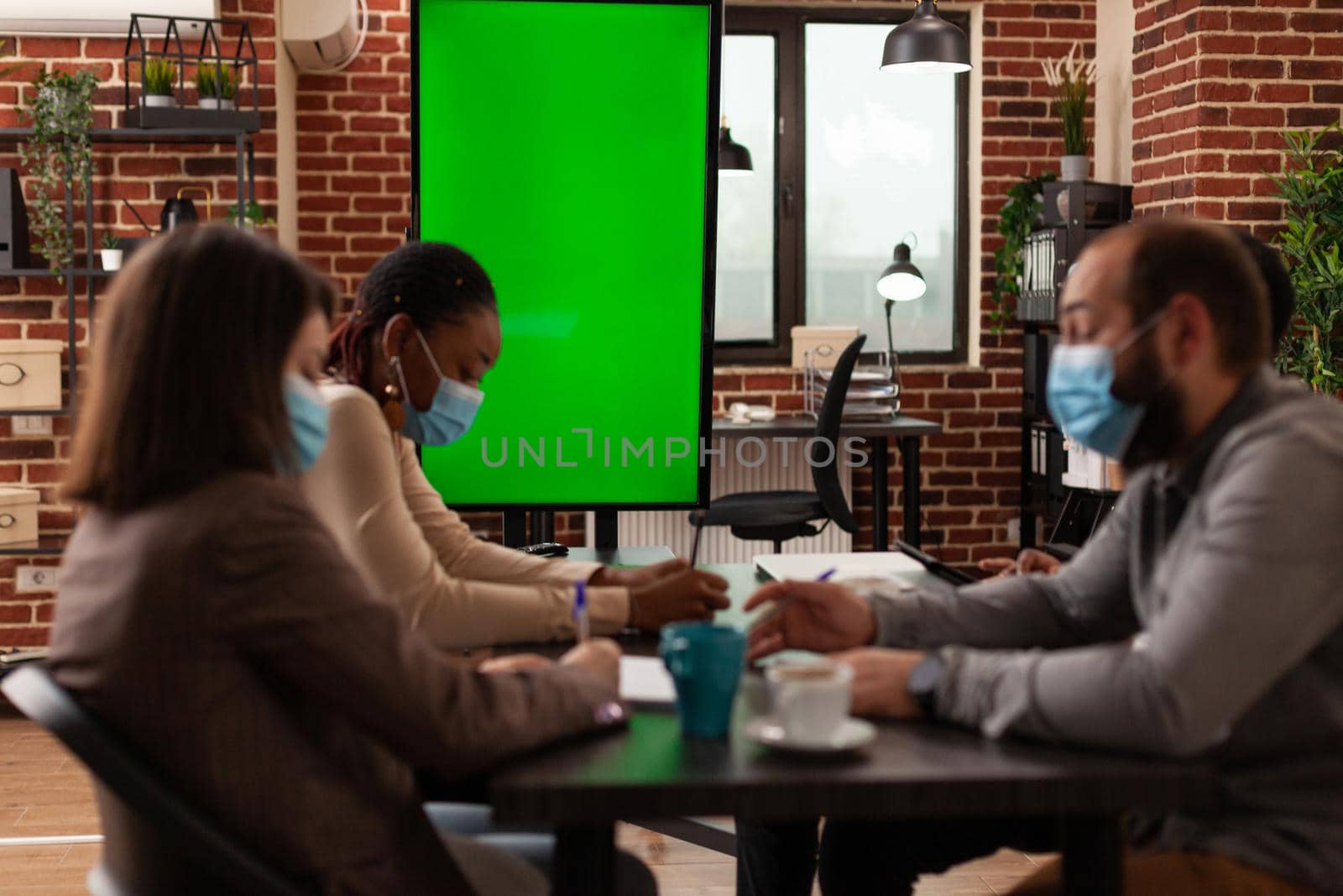 Multi-ethnic businesspeople with medical face mask against coronavirus working at business presentation in startup company meeting office. Mock up green screen chroma key monitor with isolated display