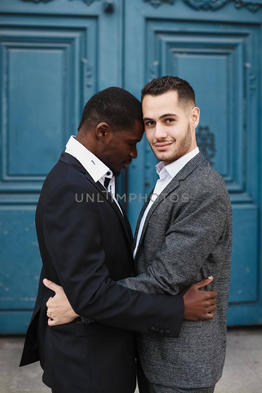 Afro american handsome man hugging caucasian guy in door background, wearing suit. Concept of same sex couple and gays.
