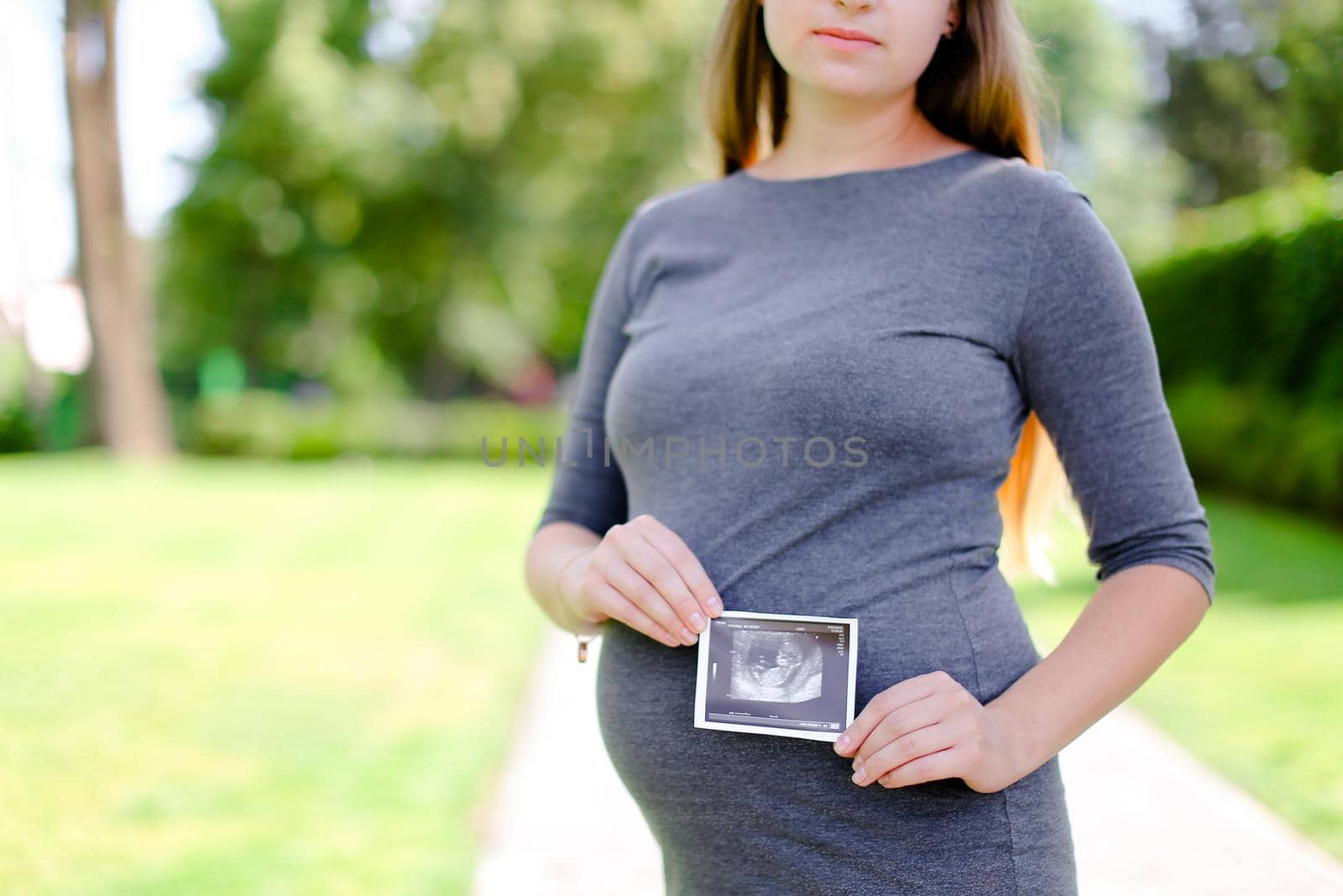 Young pregnant girl wearing grey dress keeping ultrasound photo. Concept of expectant female person and motherhood.