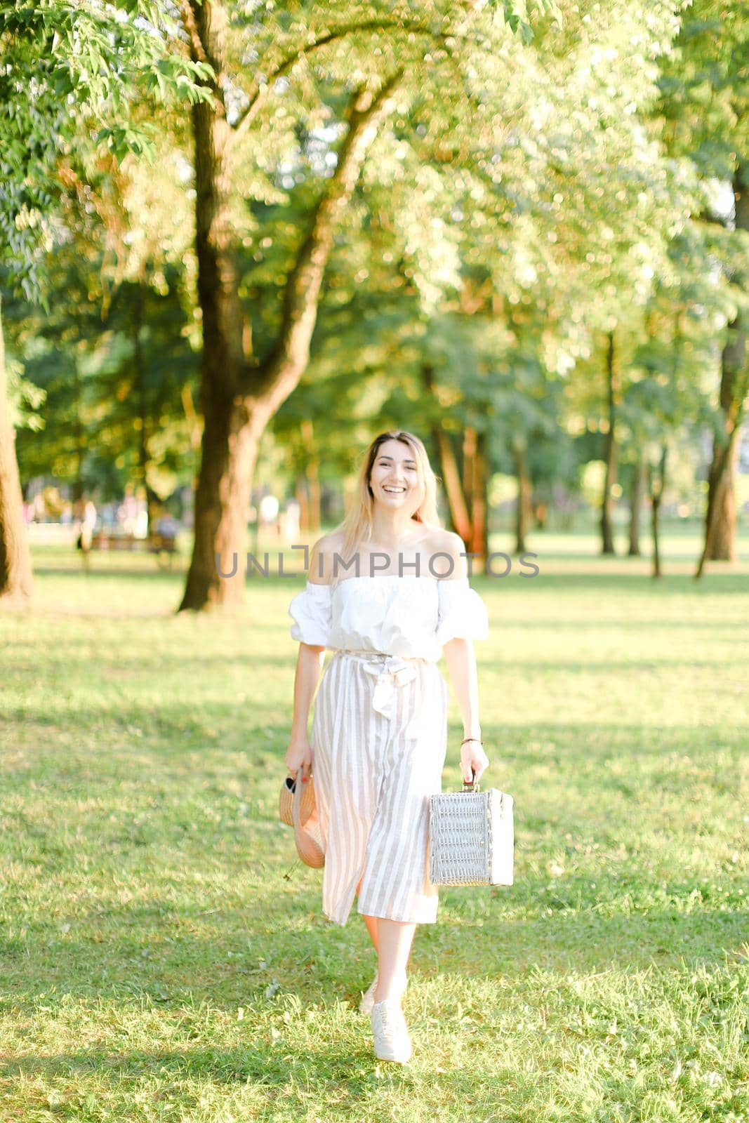 Young caucasian girl walking in garden and keeping bag, sunglasses and hat. Concept of walking in park and summer fashion.