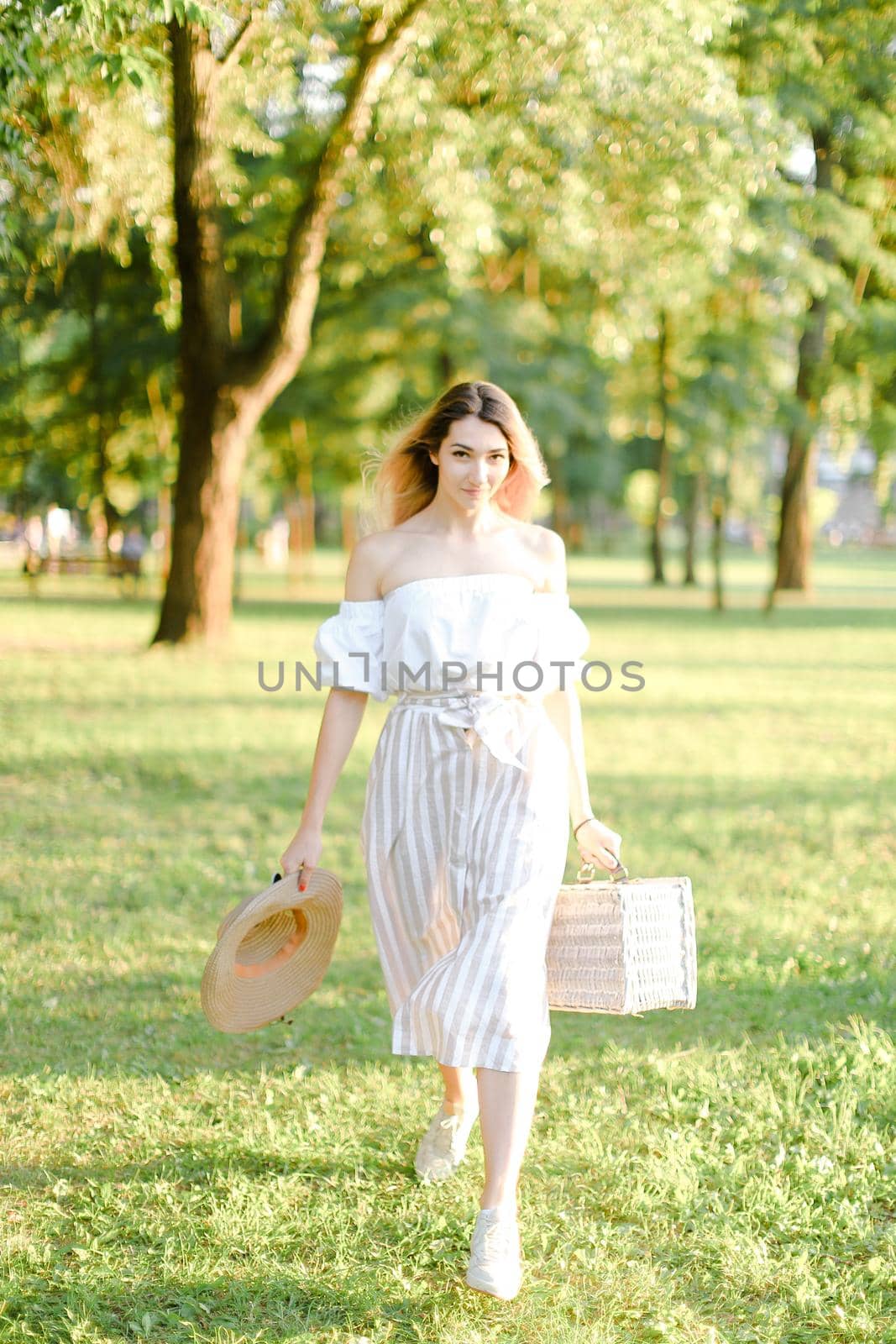 Young european woman walking in park and keeping bag, sunglasses and hat. by sisterspro