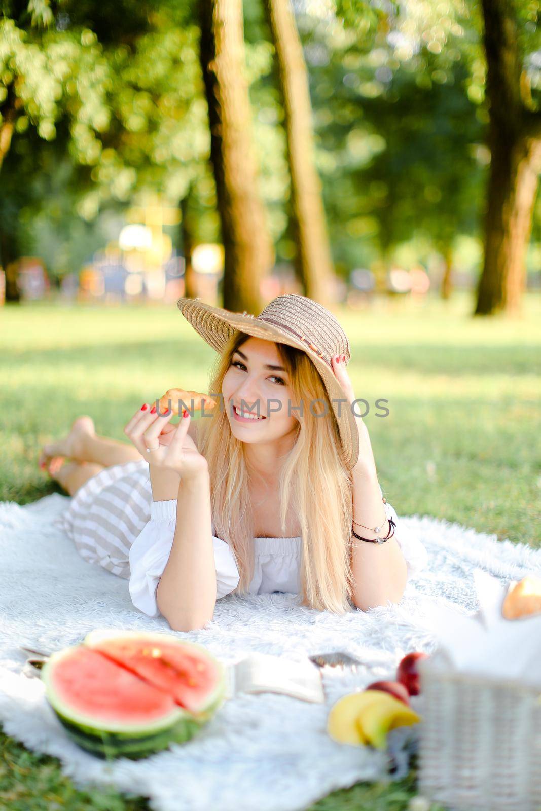 Young smiling woman in hat with croissant lying in park on plaid near watermelon. by sisterspro