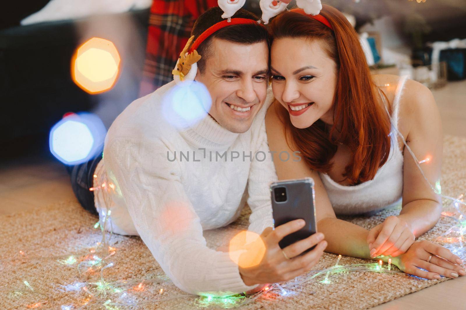 a happy married couple is lying on the floor at home near the Christmas tree and taking pictures of themselves by Lobachad