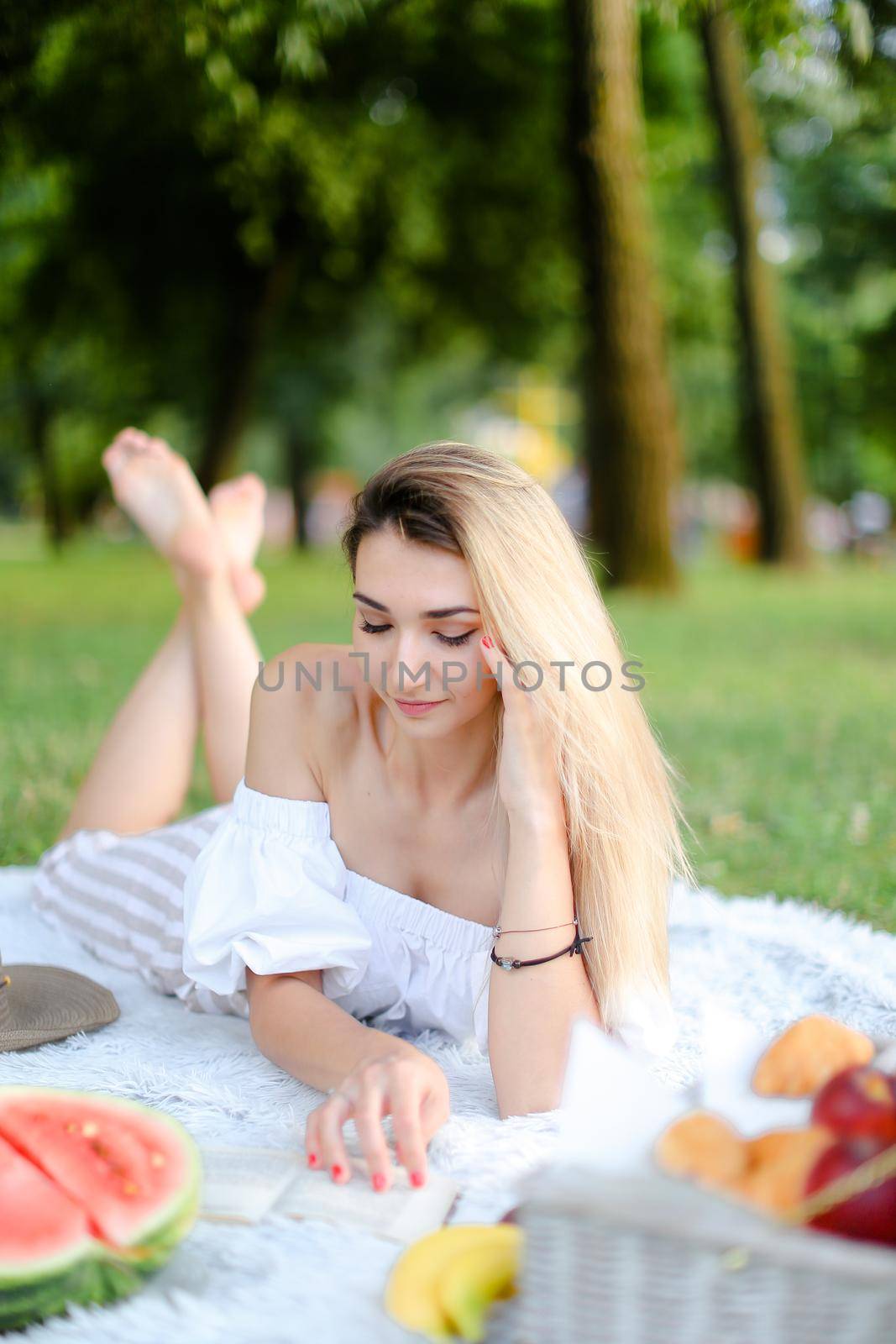 Young girl lying on plaid near watermelon in park and reading book. Concept of picnic on nature, summer vacations and leisure time.