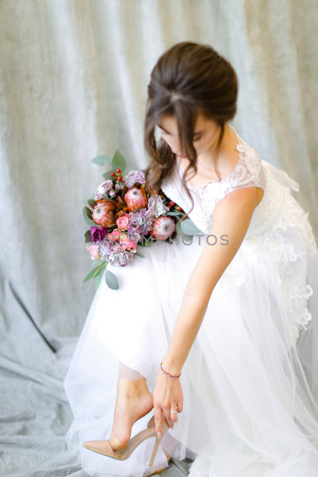 Young bride with flowers putting on shoes at photo studio. by sisterspro