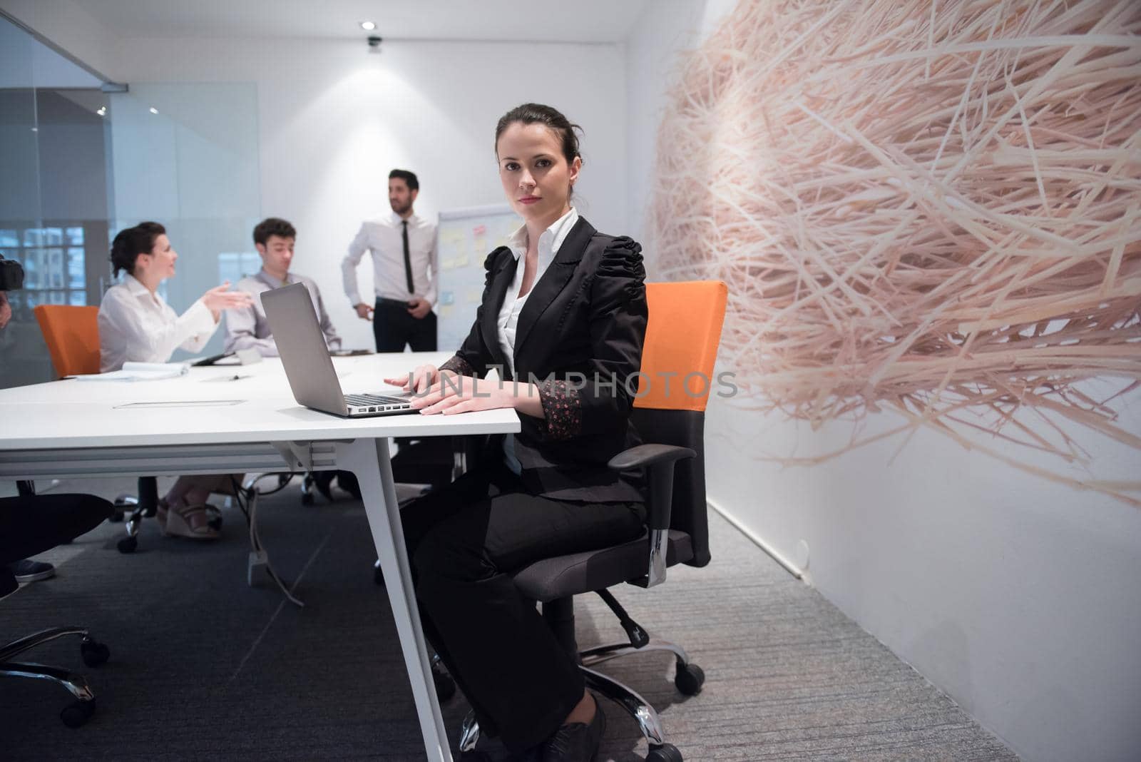 young business woman on meeting usineg laptop computer, blured group of people in background at  modern bright startup office interior taking notes on white flip board and brainstorming about plans and ideas