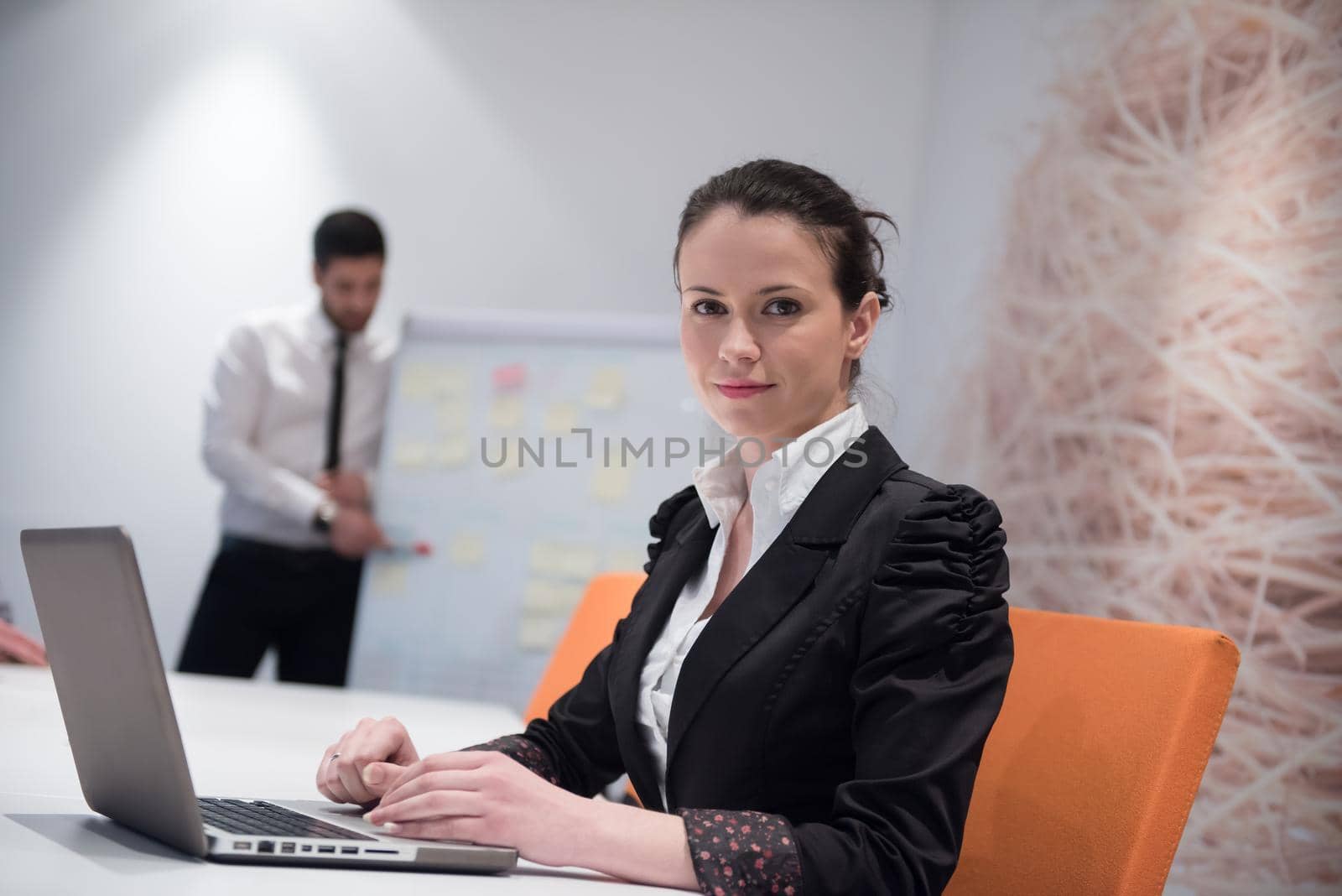 young business woman on meeting usineg laptop computer, blured group of people in background at  modern bright startup office interior taking notes on white flip board and brainstorming about plans and ideas