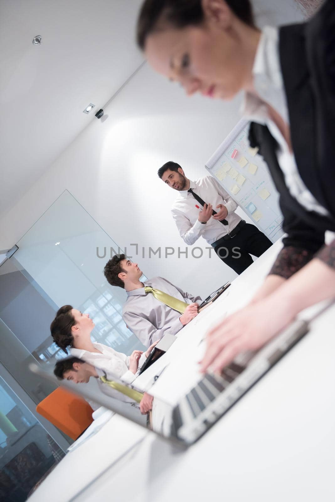 young business woman on meeting usineg laptop computer, blured group of people in background at  modern bright startup office interior taking notes on white flip board and brainstorming about plans and ideas
