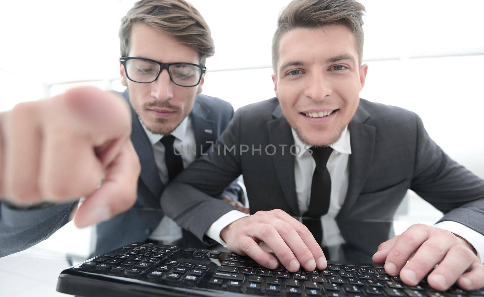 two young businessman are looking into the camera while sitting in the office. One shows the index finger