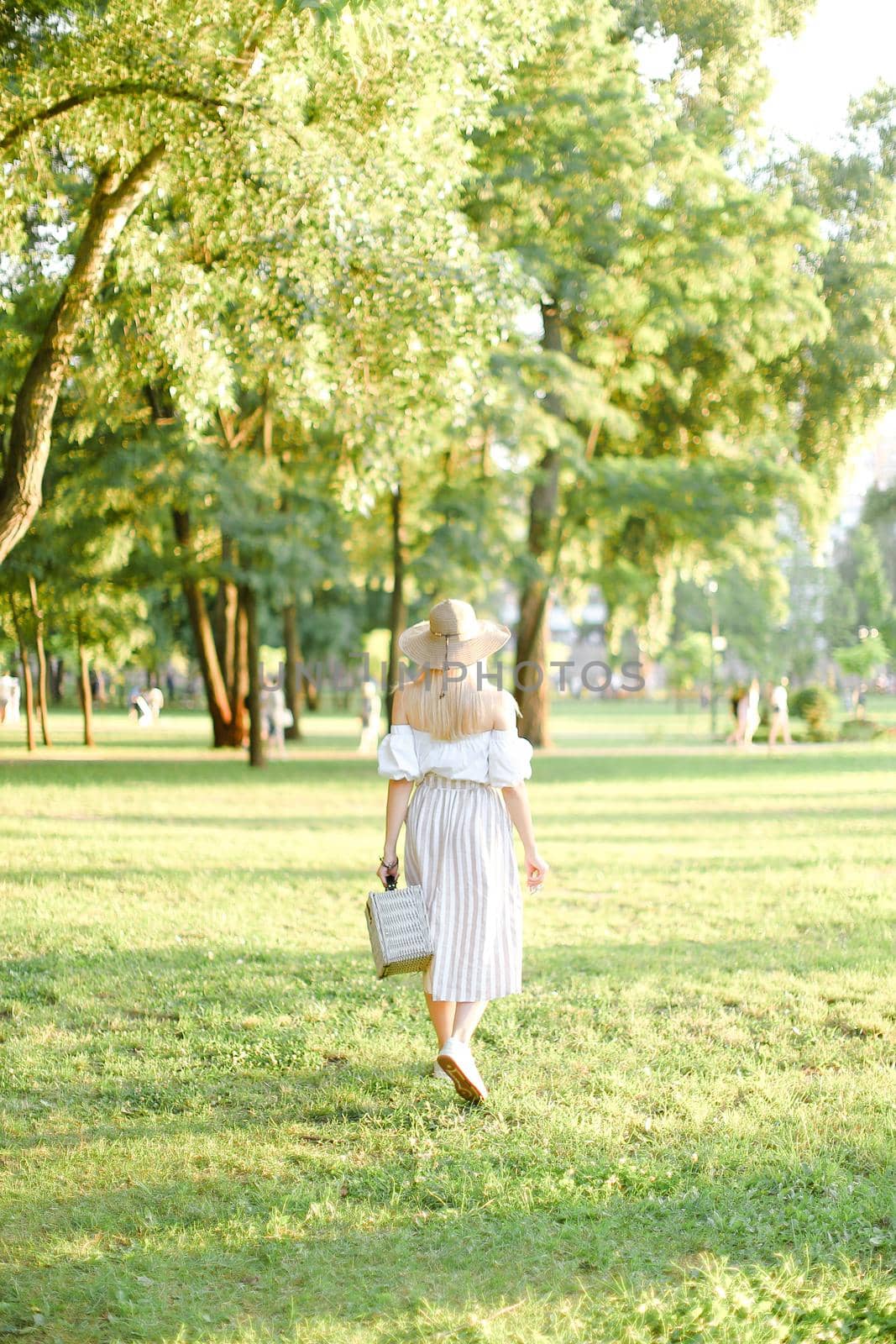 Back view of young american woman in hat walking in park and keeping bag and sunglasses. by sisterspro