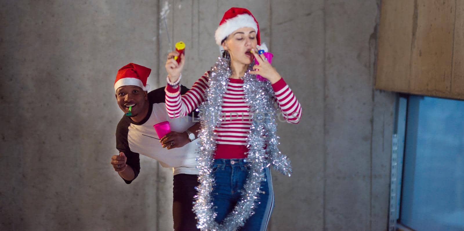 multiethnic business couple wearing red hats while celebrating new year party in new unfinished startup office with a gray wall behind them