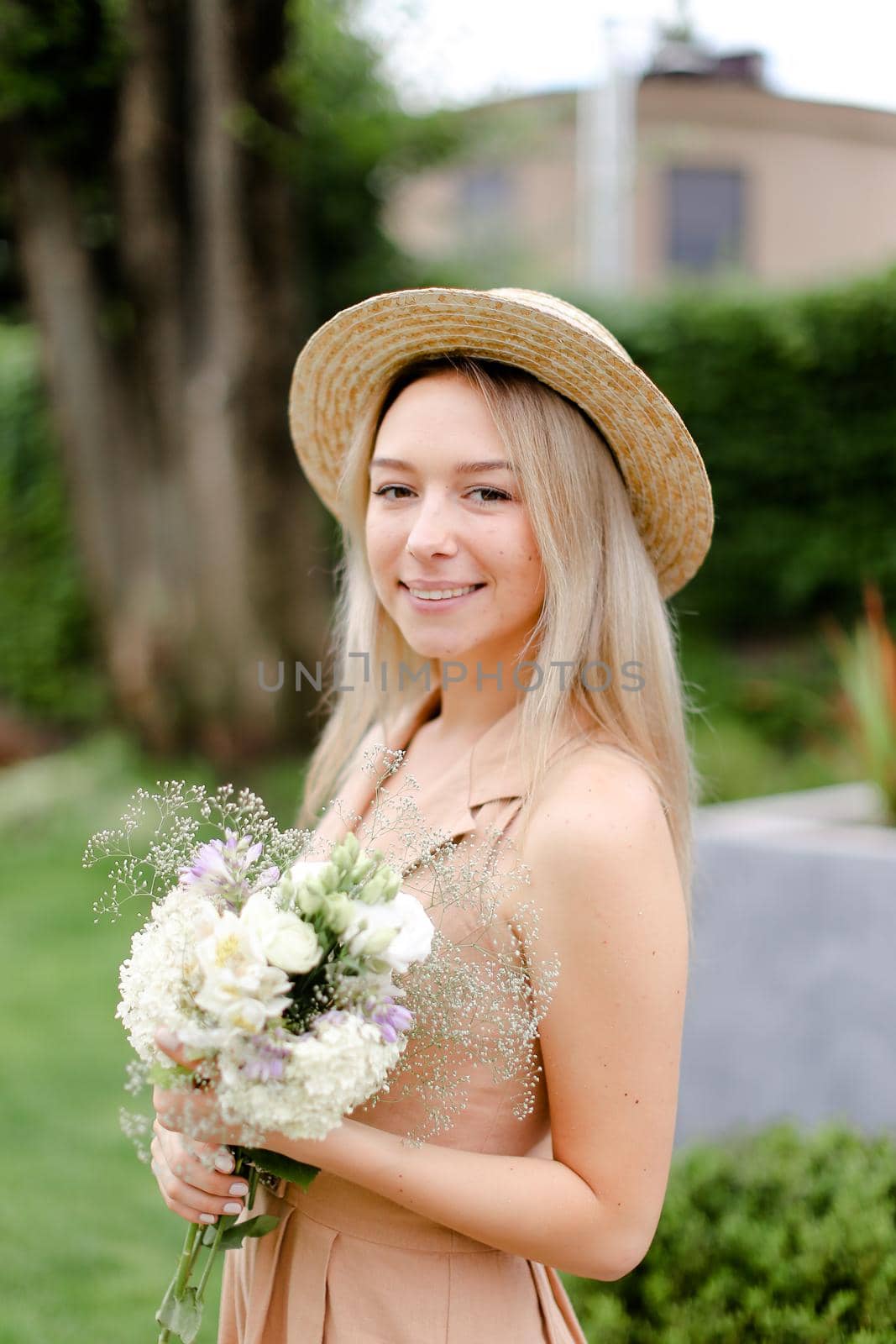Young blonde girl standing in yeard with bouquet of flowers and wearing hat with overalls. Concept of yuoth and summer season, fashion.