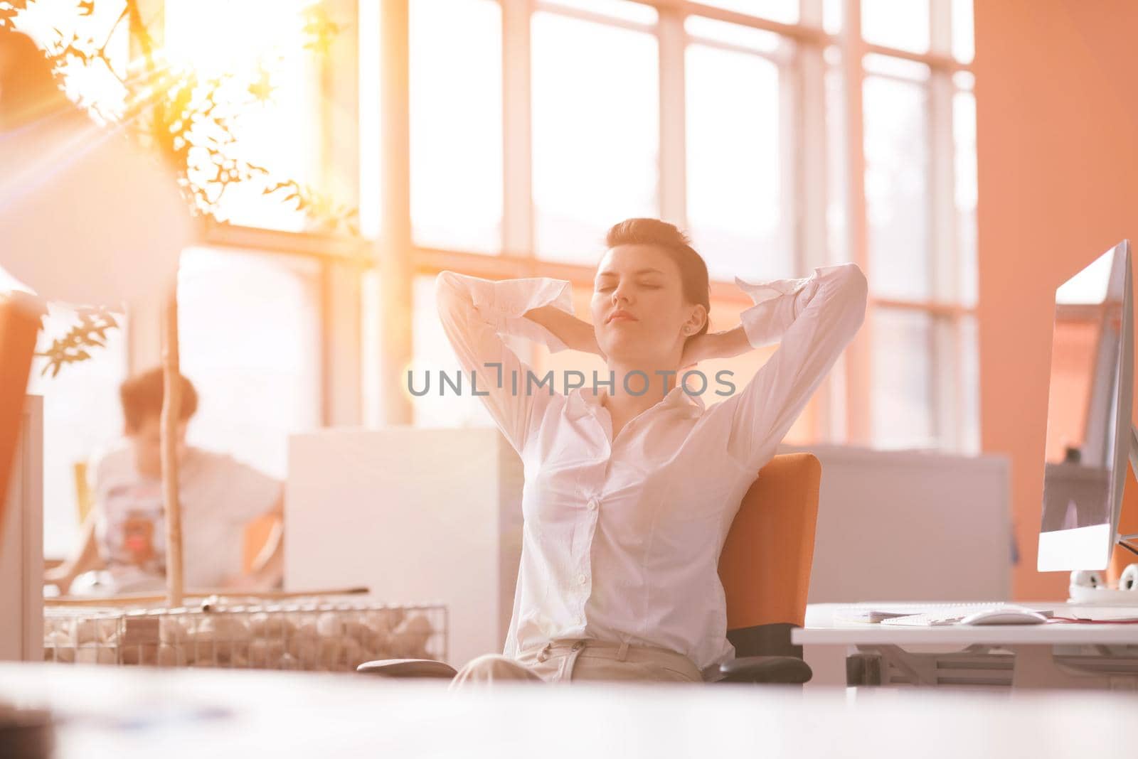 Happy young business woman relaxing and geting insiration while working on desktop computer at modern bright  starup office interior. Morning sunrise or sunset with sun flare in background.