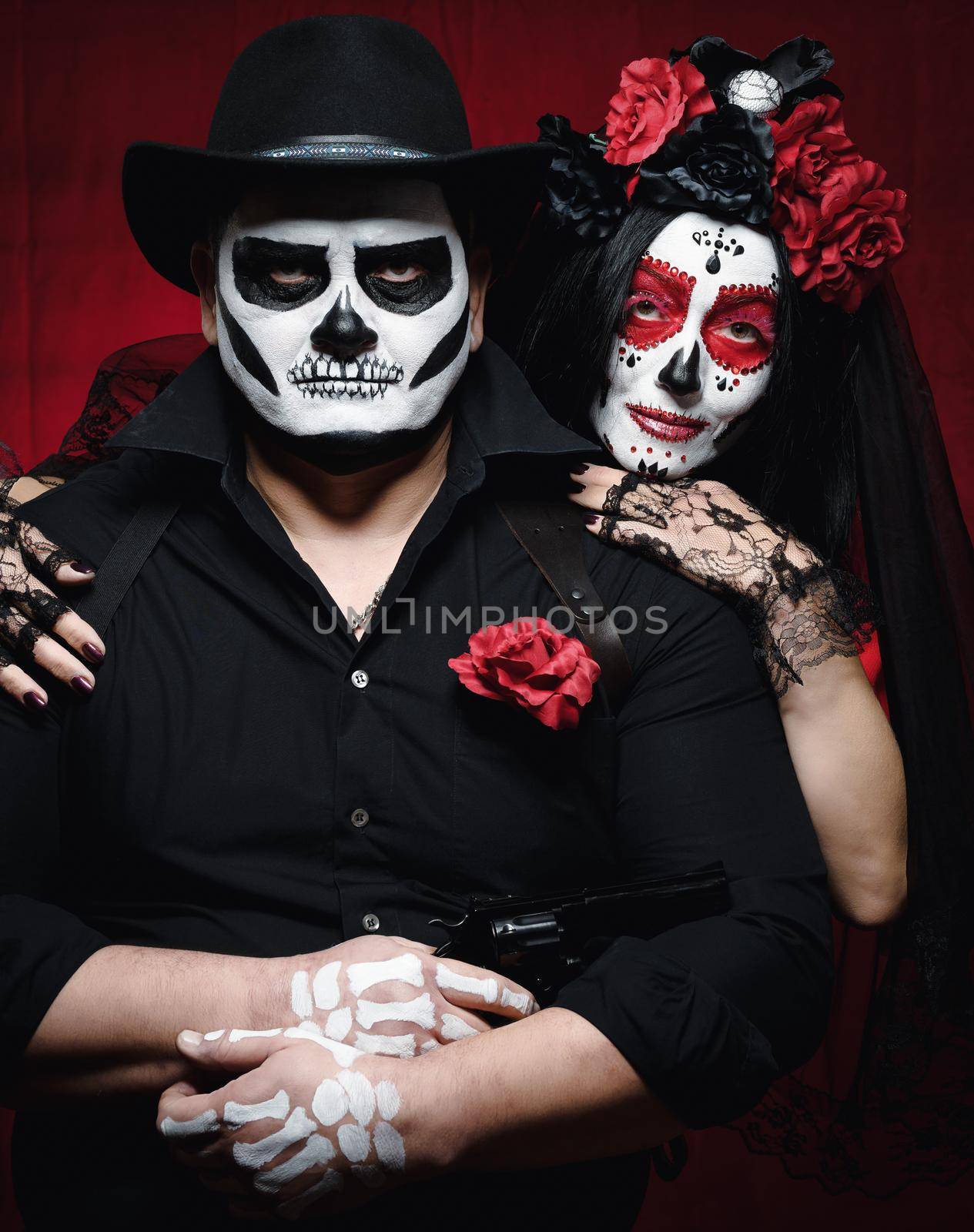 beautiful woman with a sugar skull makeup with a wreath of flowers on her head and a skeleton man in a black hat. Couple on dark red background by ndanko