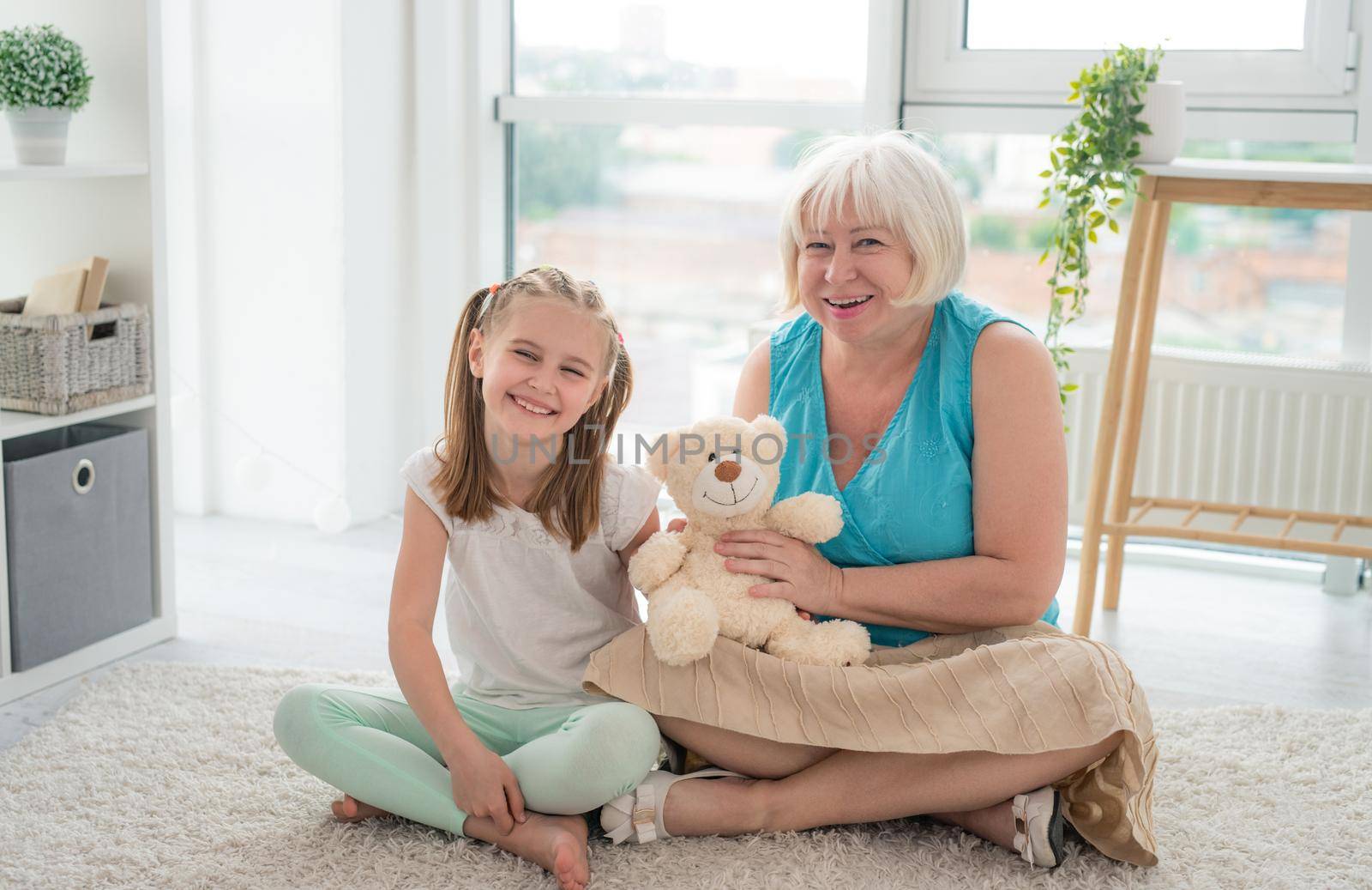 Cute little girl playing plush teddy bear toy with mature woman sitting on floor in modern apartment