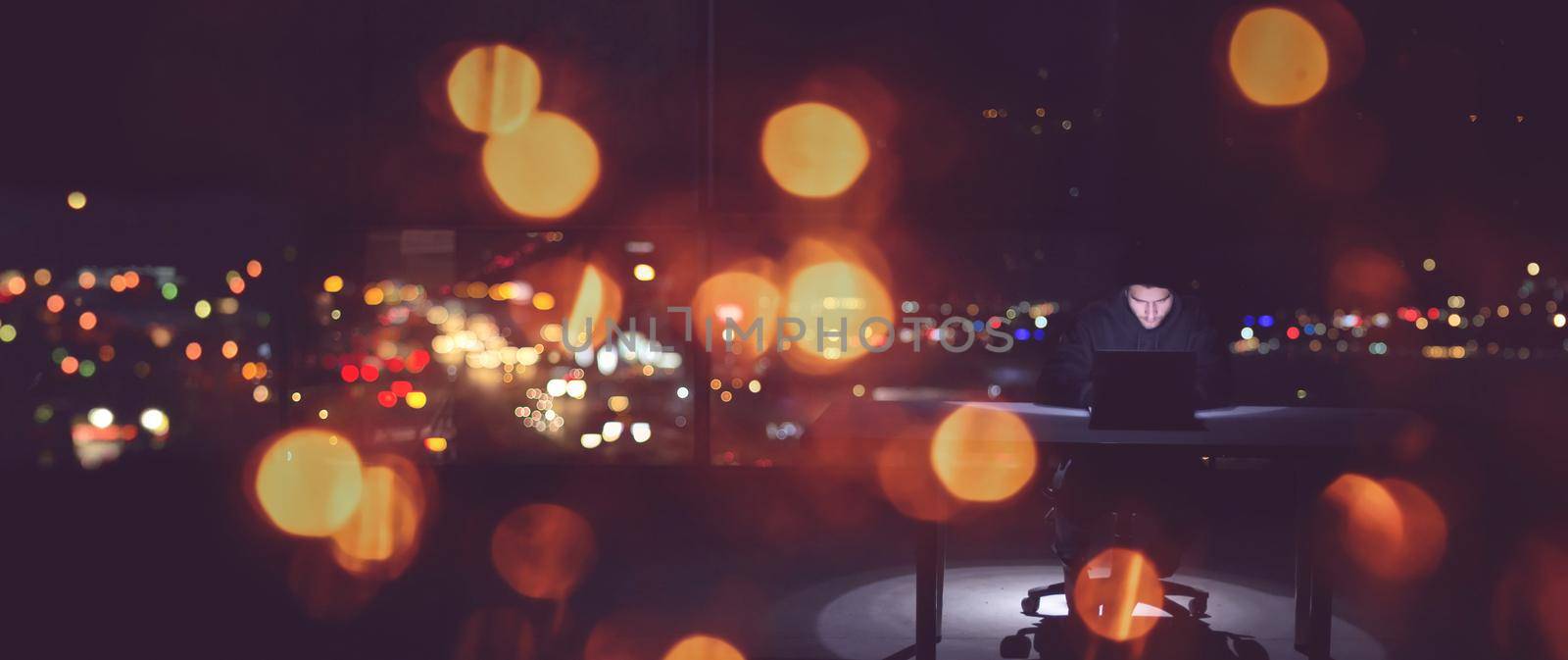 Young talented hacker using laptop computer while working in dark office with big city lights in the background at night