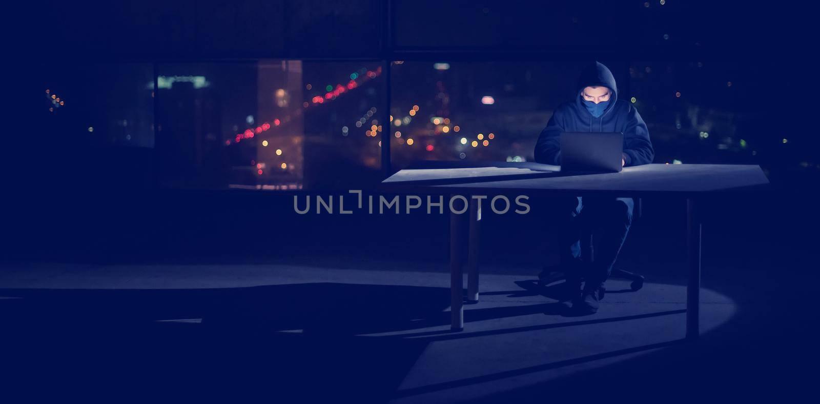 Young talented hacker using laptop computer while working in dark office with big city lights in the background at night