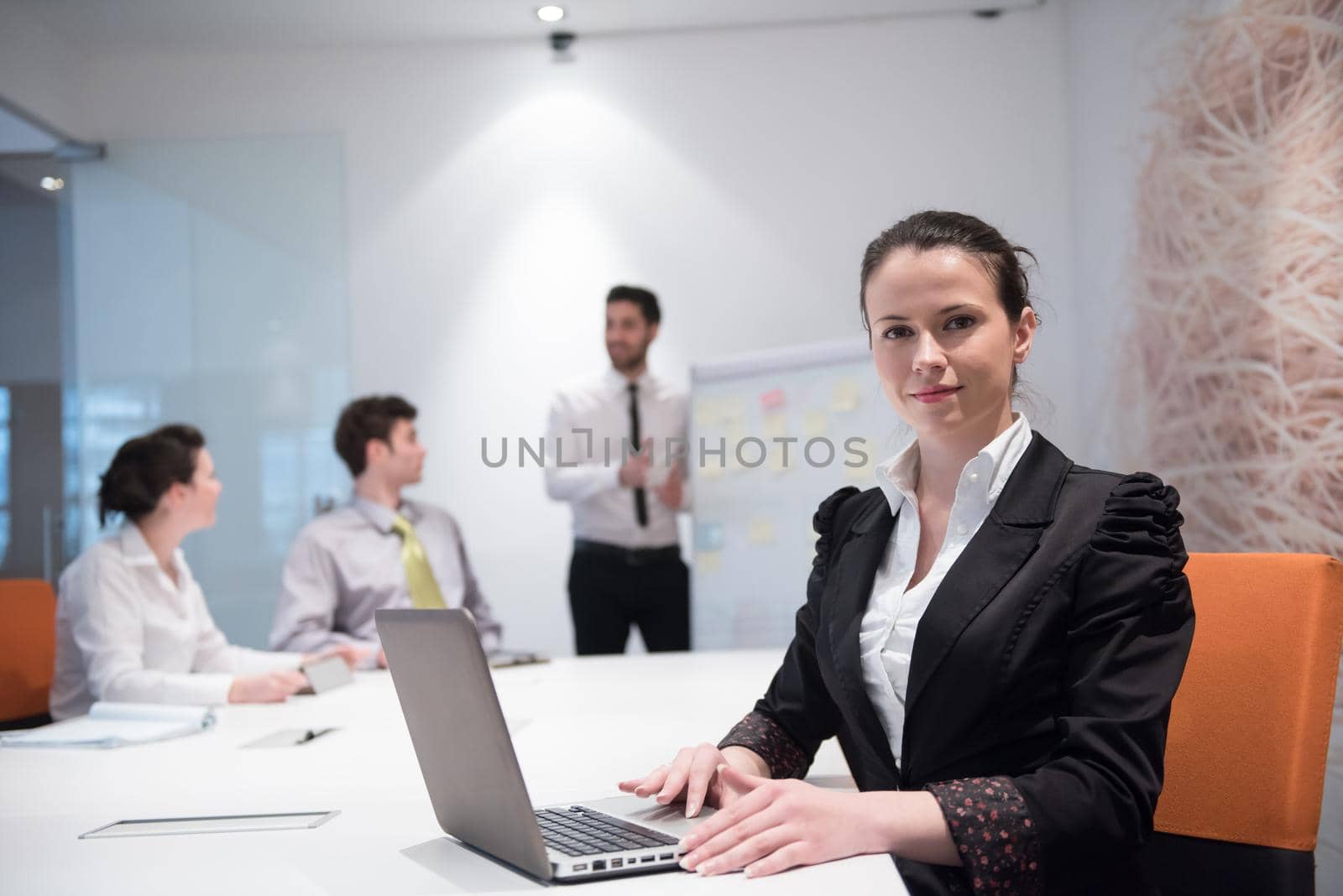 young business woman on meeting usineg laptop computer, blured group of people in background at  modern bright startup office interior taking notes on white flip board and brainstorming about plans and ideas