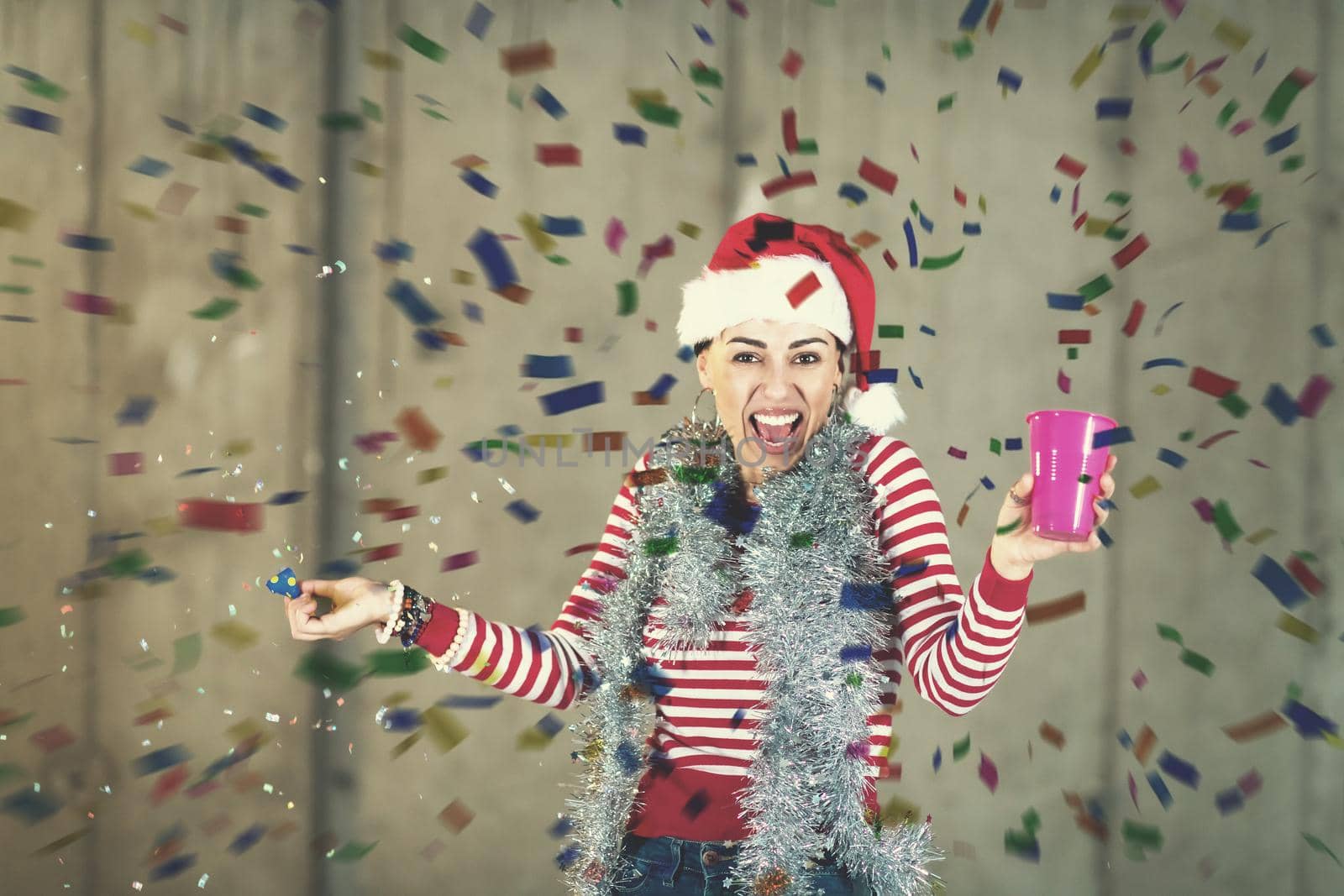 young happy casual business woman wearing a red hat and blowing party whistle while having new years confetti party in front of concrete wall