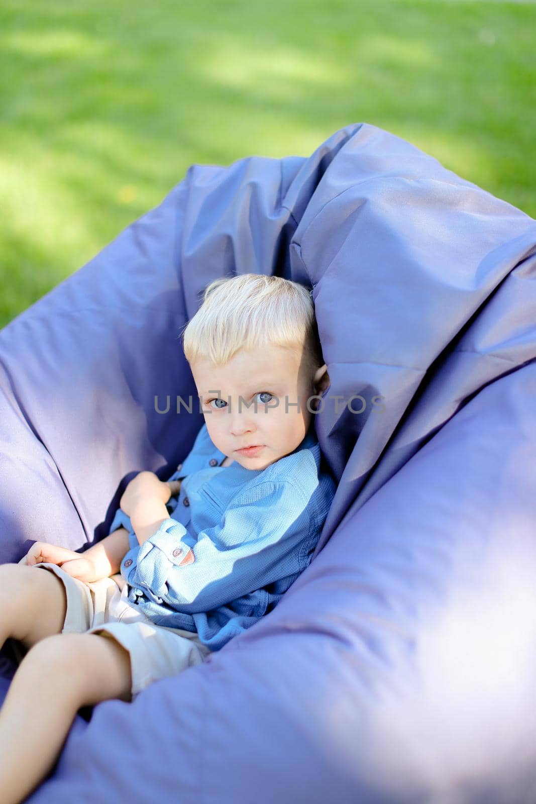 Little blond boy sitting in bag chair on grass. by sisterspro