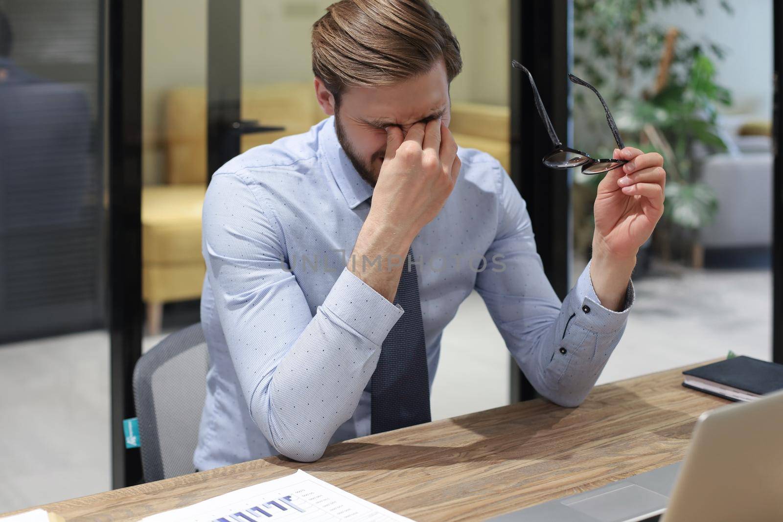 Handsome young business man with closed eyes touching face with hands while sitting on working place in office. by tsyhun