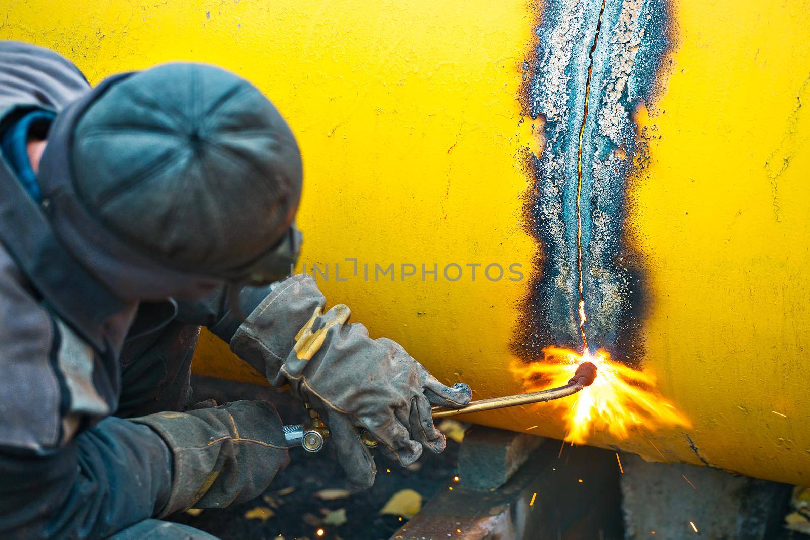The welder cuts a large yellow pipe with acetylene welding for gasification. Disposal of old used metal pipes. Authentic workflow scene. Industrial background.