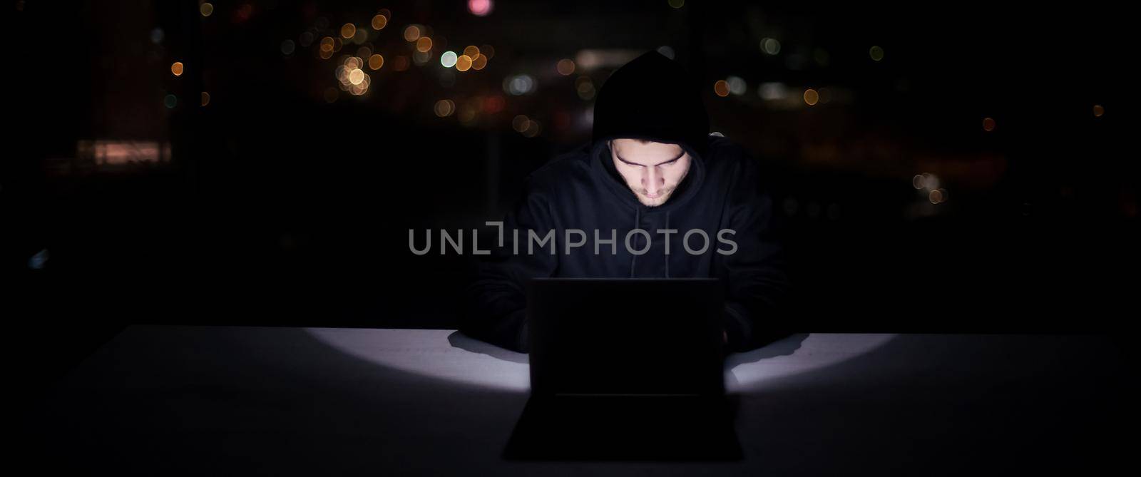 Young talented hacker using laptop computer while working in dark office with big city lights in the background at night