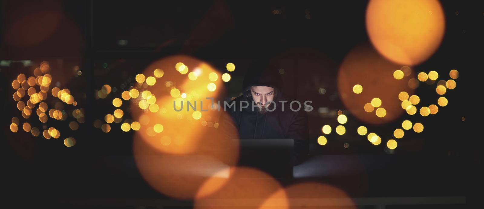 Young talented hacker using laptop computer while working in dark office with big city lights in the background at night