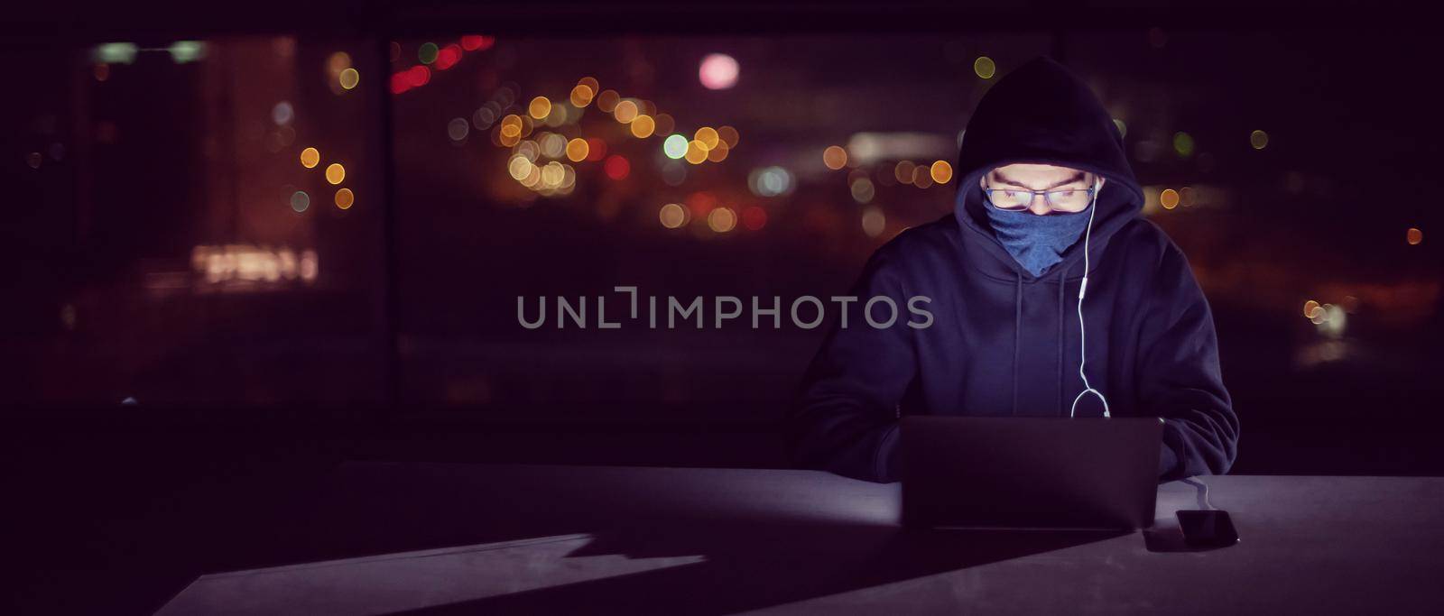 Young talented hacker using laptop computer while working in dark office with big city lights in the background at night