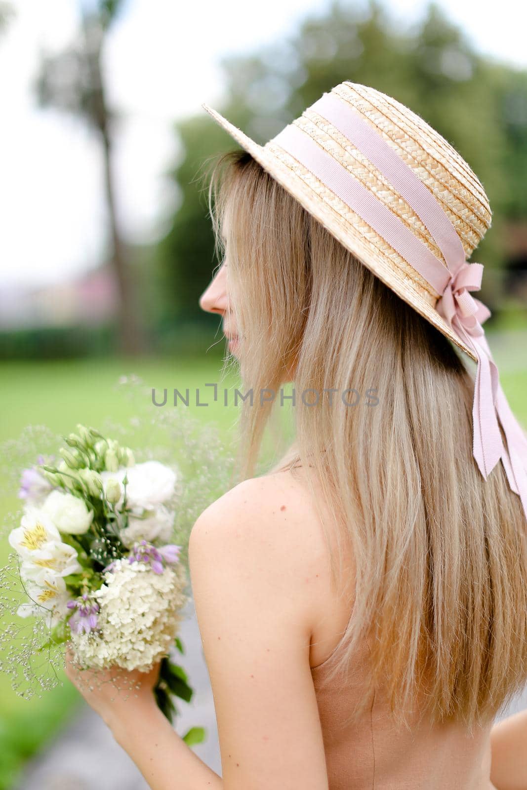 Back view of young blonde woman in body color overalls and hat standing in yeard with flowers. Concept of fashion and summer season.