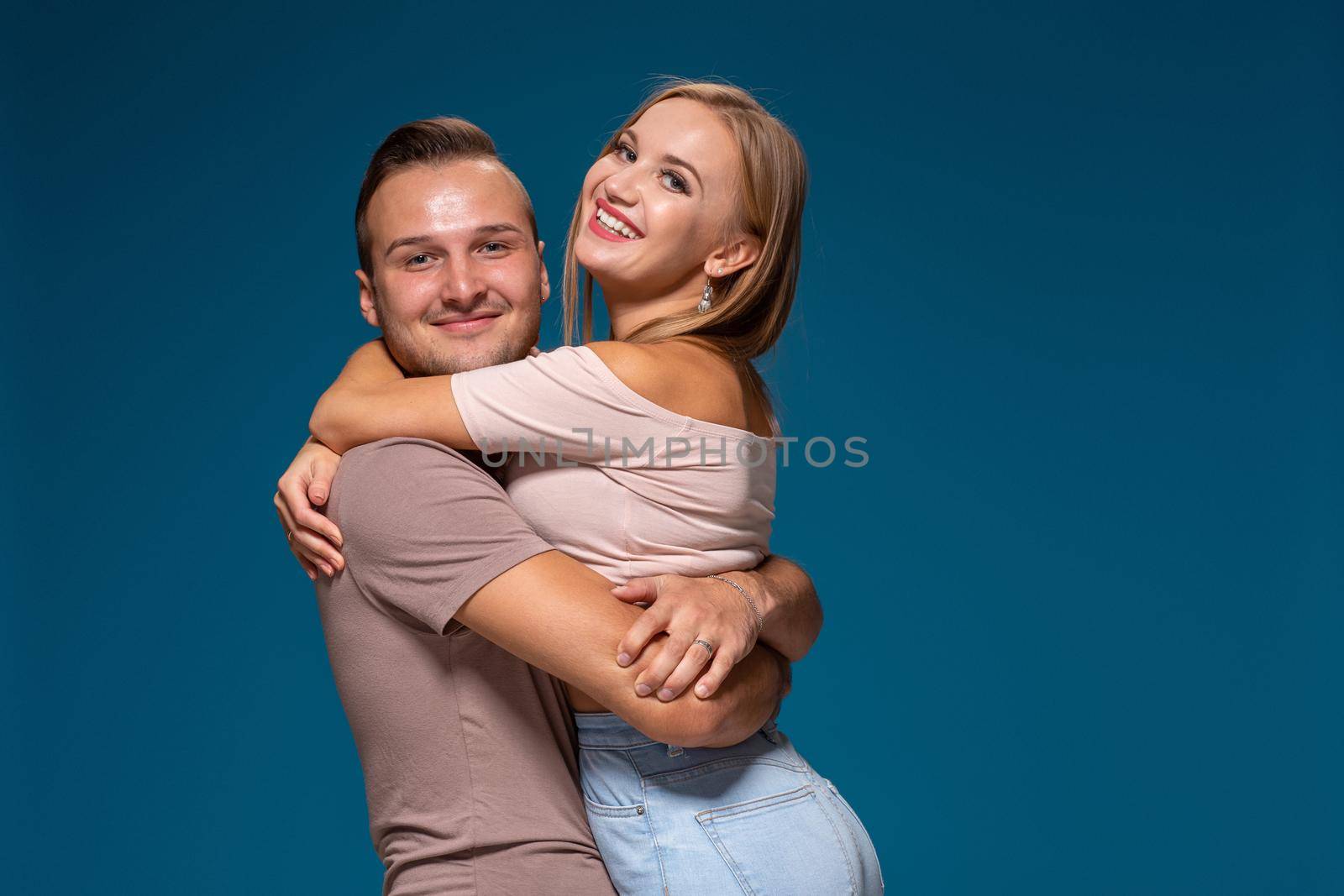 Young couple is hugging on blue background in studio. They wear T-shirts, jeans and smile. Friendship, love and relationships concept